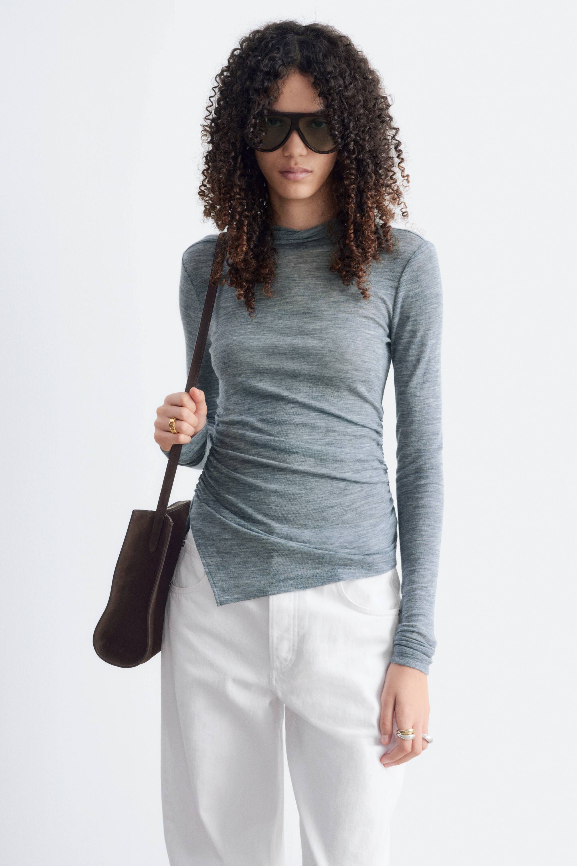 Grotere afbeelding bekijken: De model draagt een slim-fit top in grijze melange woljersey met lange mouwen, een opstaande kraag en geplooide details aan de taille die overgaan in een asymmetrische zoom. De top is gestyled met een witte broek en het model draagt een donkerbruine schoudertas.