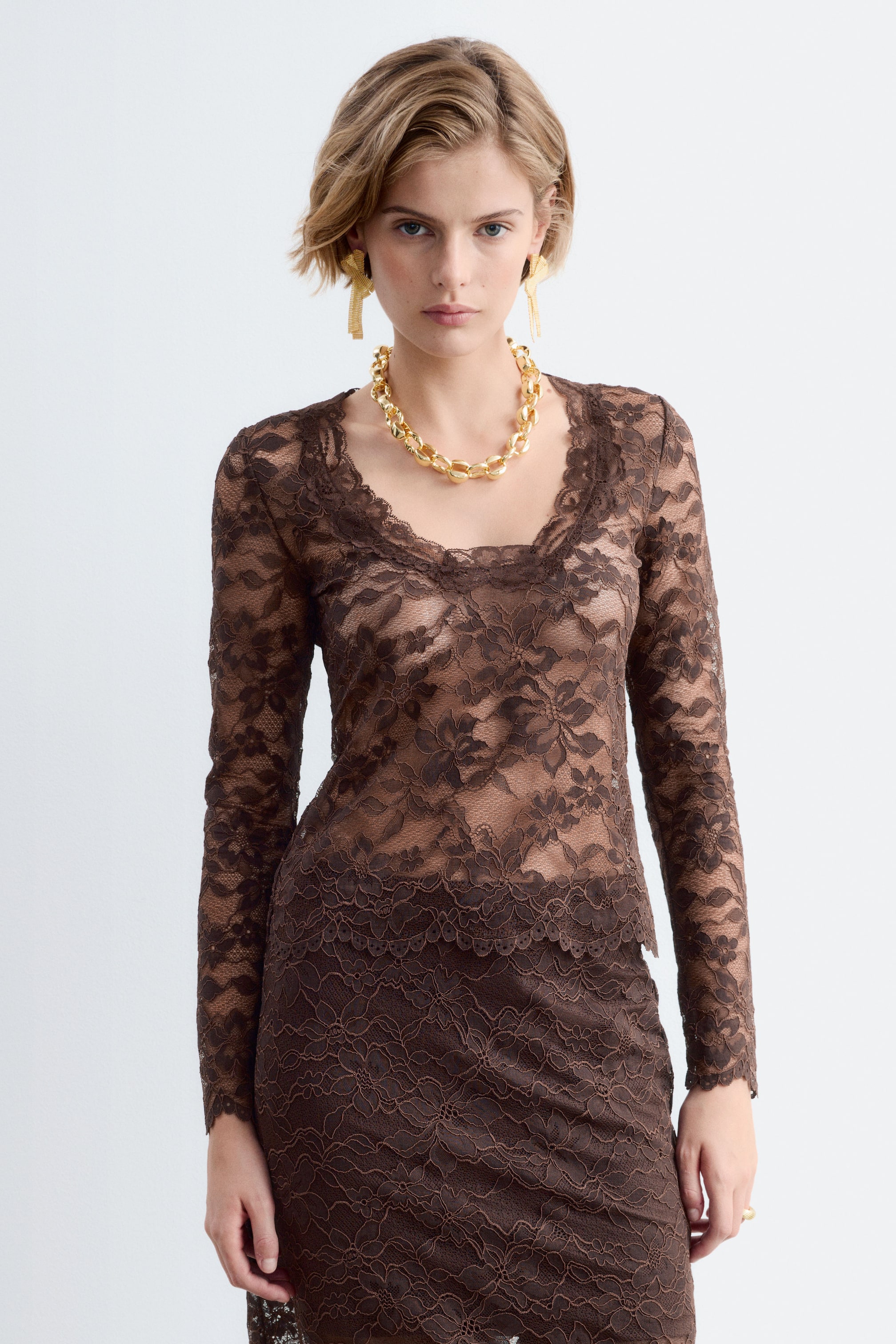 Vezi imaginea mai mare: Un top maro din dantelă, semi-transparent, cu mâneci lungi, decolteu rotund și margini festonate, este purtat de model. Topul slim-fit este stilizat cu o fustă asortată din dantelă maro și un colier gros cu lanț auriu.