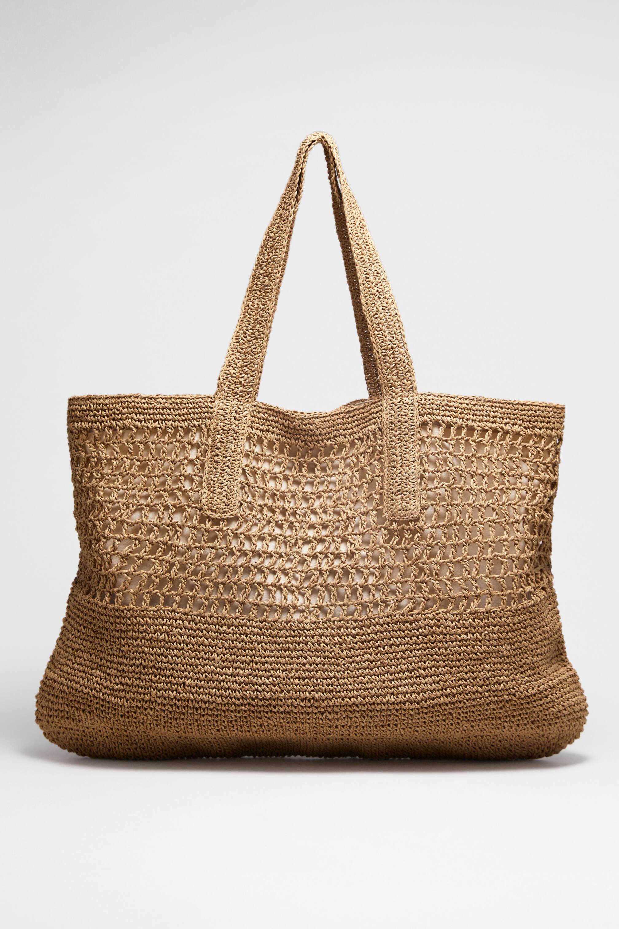 Agrandir l'image: Un sac fourre-tout tissé de couleur naturelle est mis en avant, avec un tissage ouvert en forme de filet sur sa partie supérieure et un tissage plus serré et solide sur sa partie inférieure, ainsi que deux poignées supérieures. Une doublure de couleur claire est visible à travers le tissage ouvert du sac.