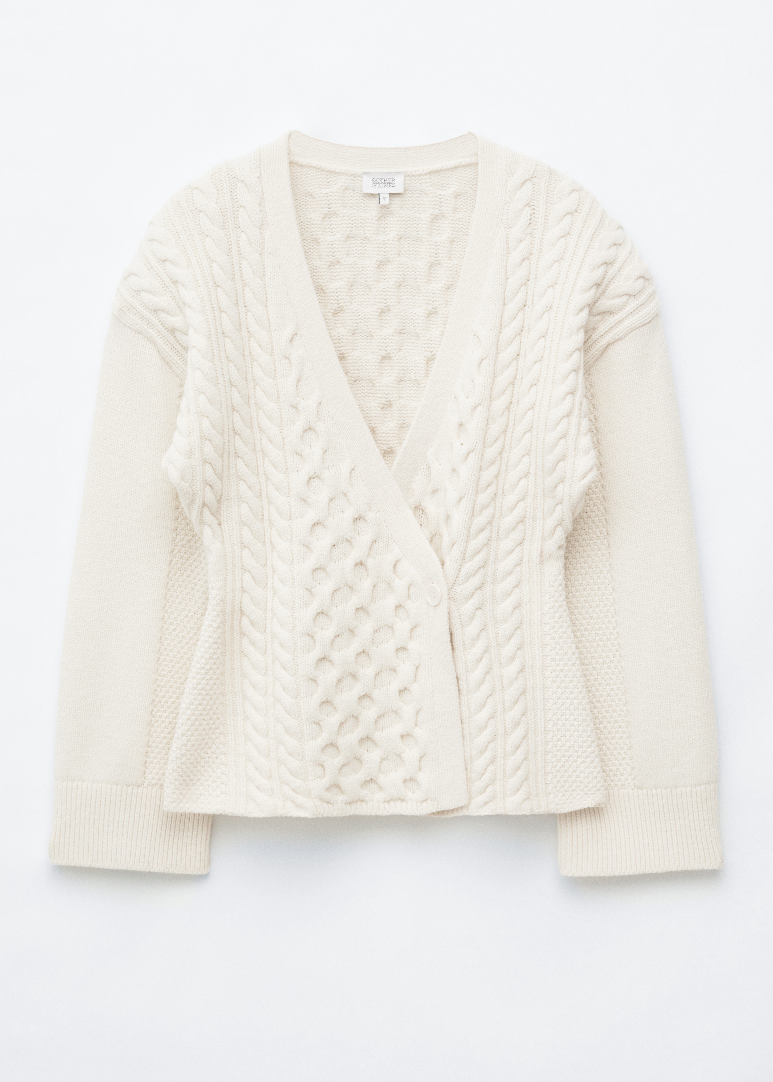 Un cardigan enveloppant en maille torsadée crème est porté, affichant un décolleté en V profond et une fermeture à un seul bouton à la taille. Il présente divers motifs de maille torsadée, y compris des motifs à boules et torsadés, avec des poignets et un ourlet côtelés, tandis qu'une sangle de sac sombre traverse son épaule gauche.
