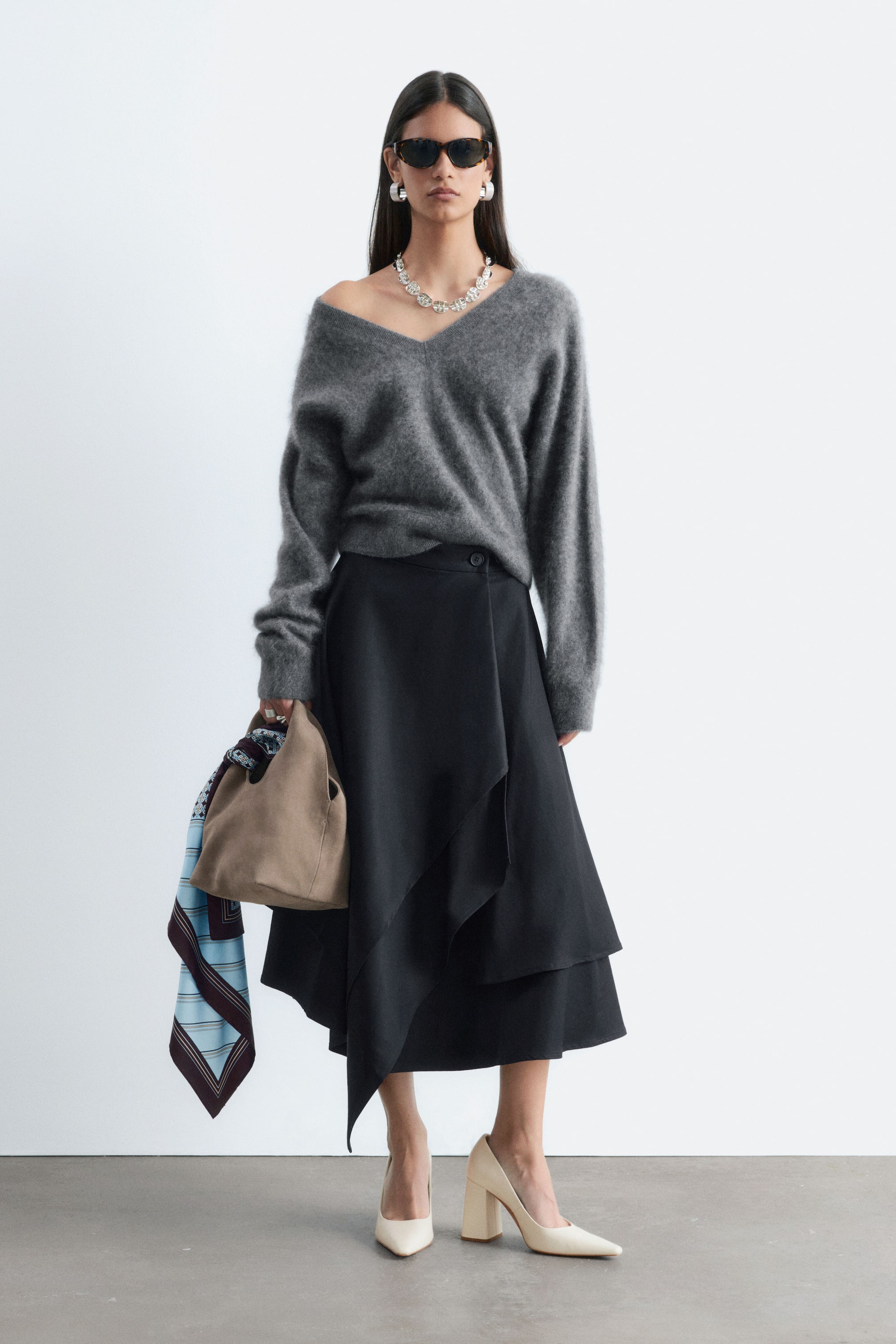 Grotere afbeelding bekijken: Een zwarte midi-rok met dubbele laag en een asymmetrische, gedrapeerde snit wordt gedragen door het model en gestyled met een grijze gebreide trui die ingestopt is. De rok heeft een knoopsluiting in de taille en een vloeiende, gelaagde zoomlijn.