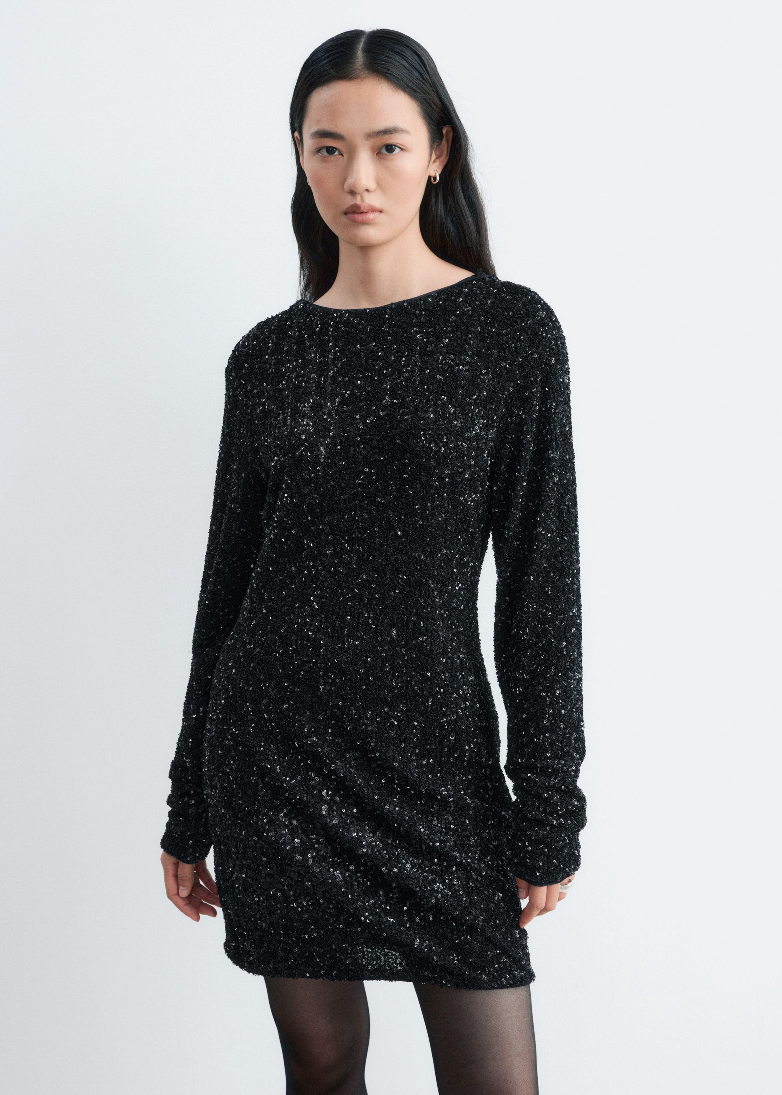 The model is wearing a black sequined long-sleeve mini dress, standing and facing forward. The dress features a crew neckline and is styled with sheer black tights.