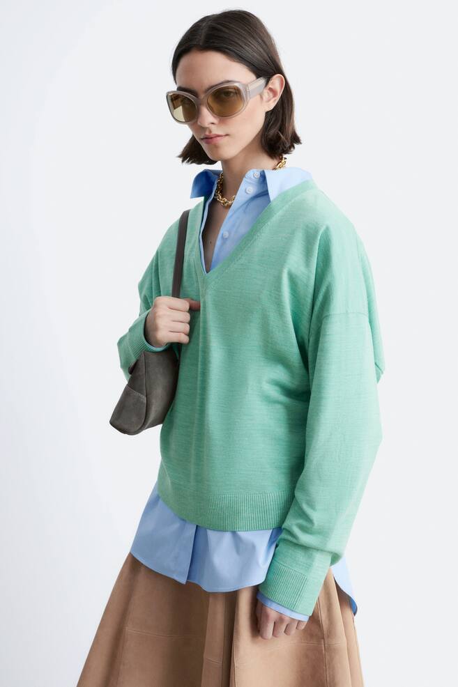 A light green fine knit V-neck jumper is showcased from a front-right angle, featuring a relaxed fit, drop shoulders, and ribbed cuffs and hem. It is being worn layered over a light blue collared shirt, highlighting its V-neckline. - 1