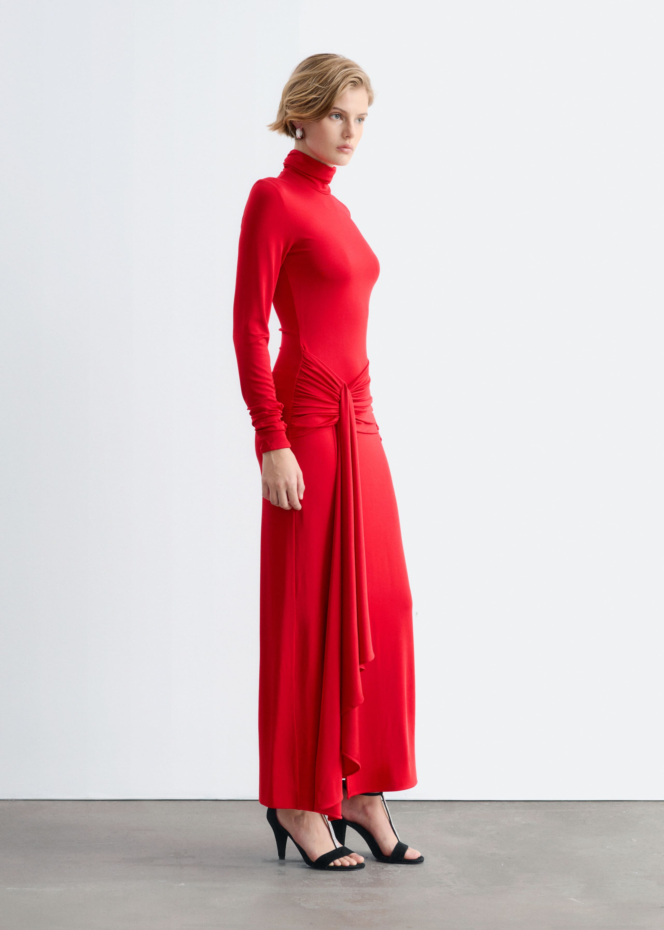 The model is wearing a red jersey maxi dress with a mock neck, long sleeves and a draped tie detail at the waist. The slim-fitting dress falls to the ankles and is styled with black high-heeled sandals.
