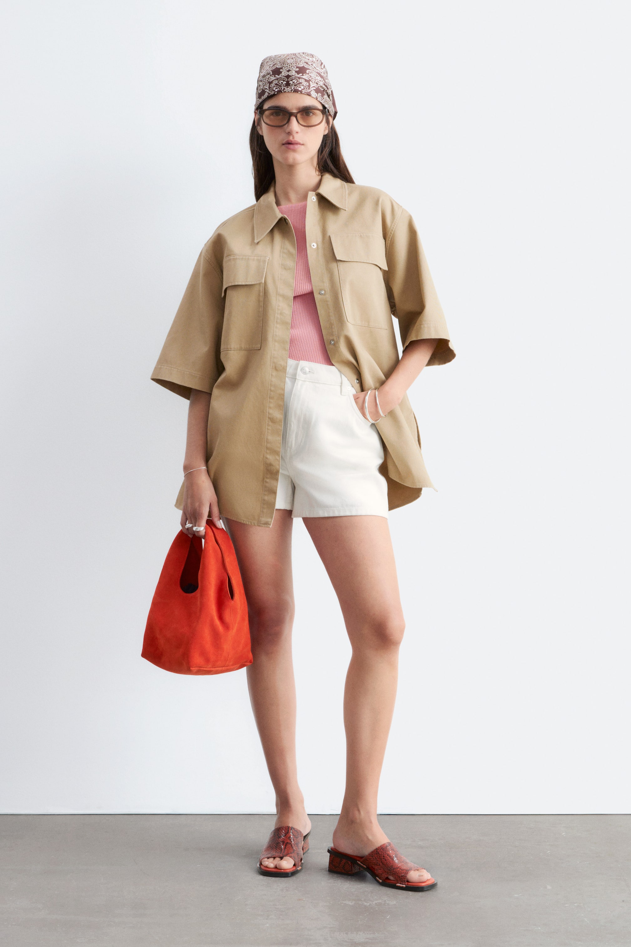 Agrandir l'image: Une chemise utilitaire beige est présentée de face, avec une coupe ample et décontractée, ainsi que des manches courtes et larges. Elle est conçue avec un col pointu, deux grandes poches à rabat sur la poitrine, et une fermeture à boutons sur le devant, portée ouverte sur un haut rose.
