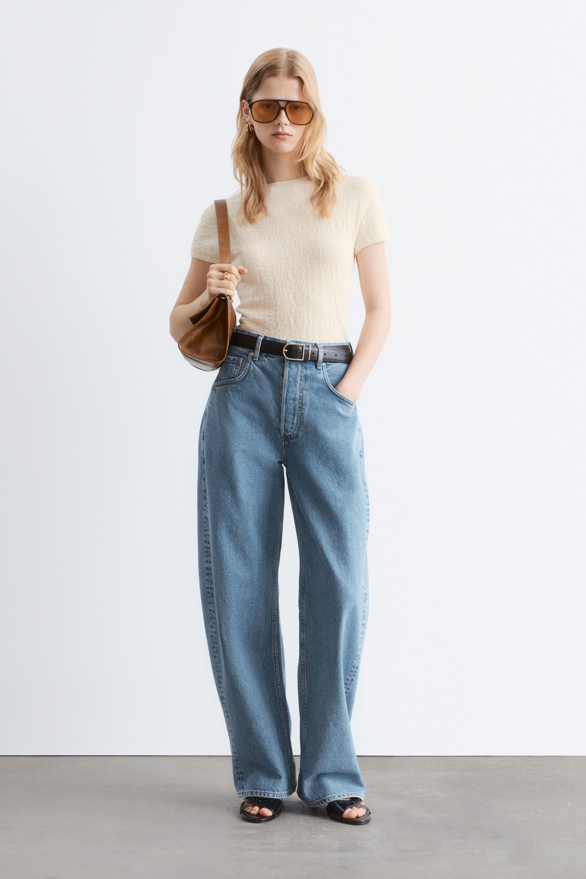Visualizza immagine più grande: Un outfit presenta un top a maniche corte in maglia testurizzata color crema con scollo tondo, infilato in jeans a vita alta blu a gamba larga, e una cintura in pelle nera con fibbia rettangolare, il tutto presentato frontalmente. Il look è accessoriato con occhiali da sole aviator marroni, una piccola borsa a tracolla in pelle marrone, sandali neri aperti in punta, orecchini a cerchio in oro e un anello in oro.