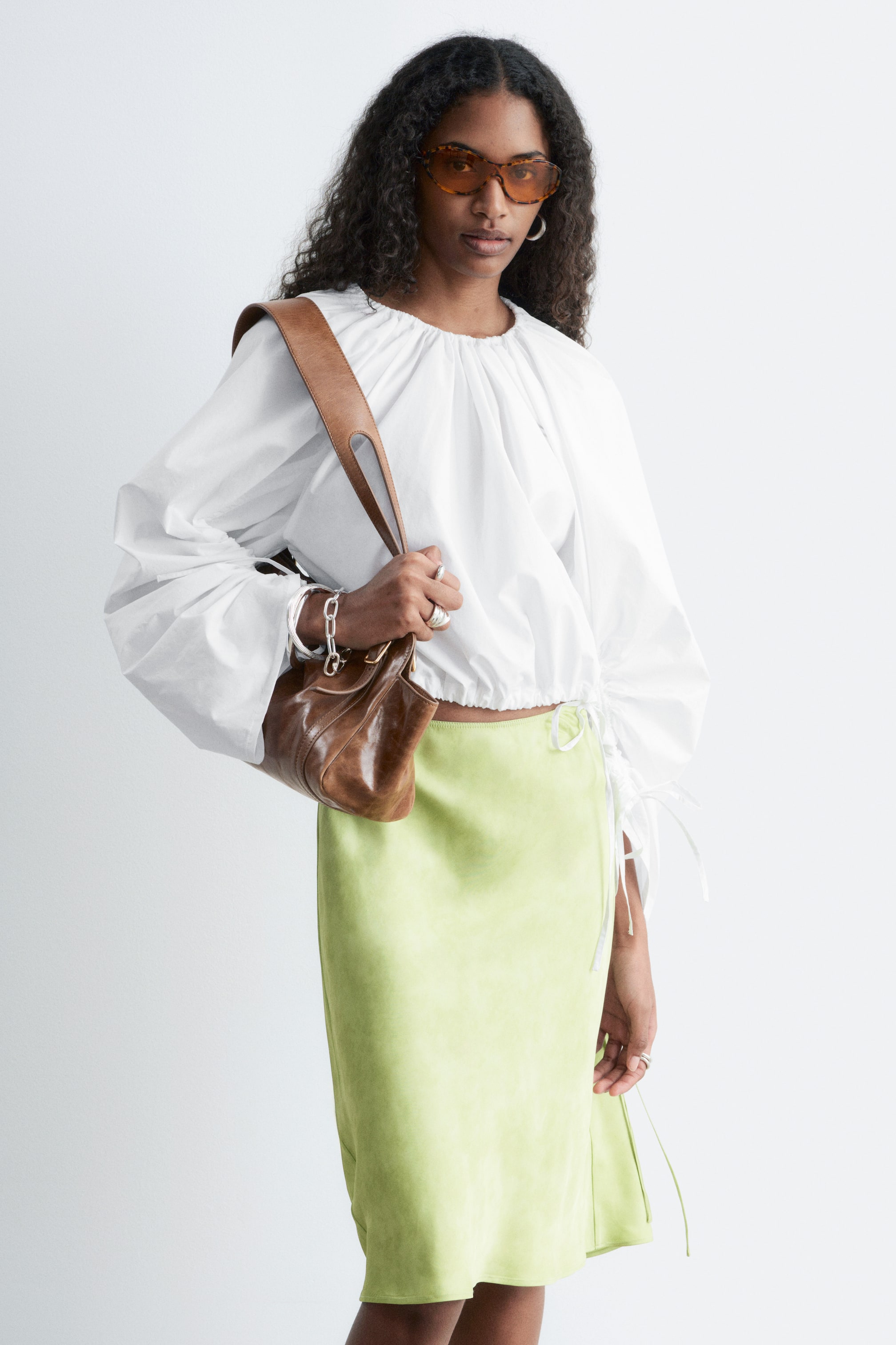 Visualizza immagine più grande: Una blusa scultorea bianca è indossata dalla modella, caratterizzata da uno scollo arricciato e maniche voluminose. Lacci a coulisse stringono l'orlo corto e i polsini delle maniche, creando una texture arricciata.