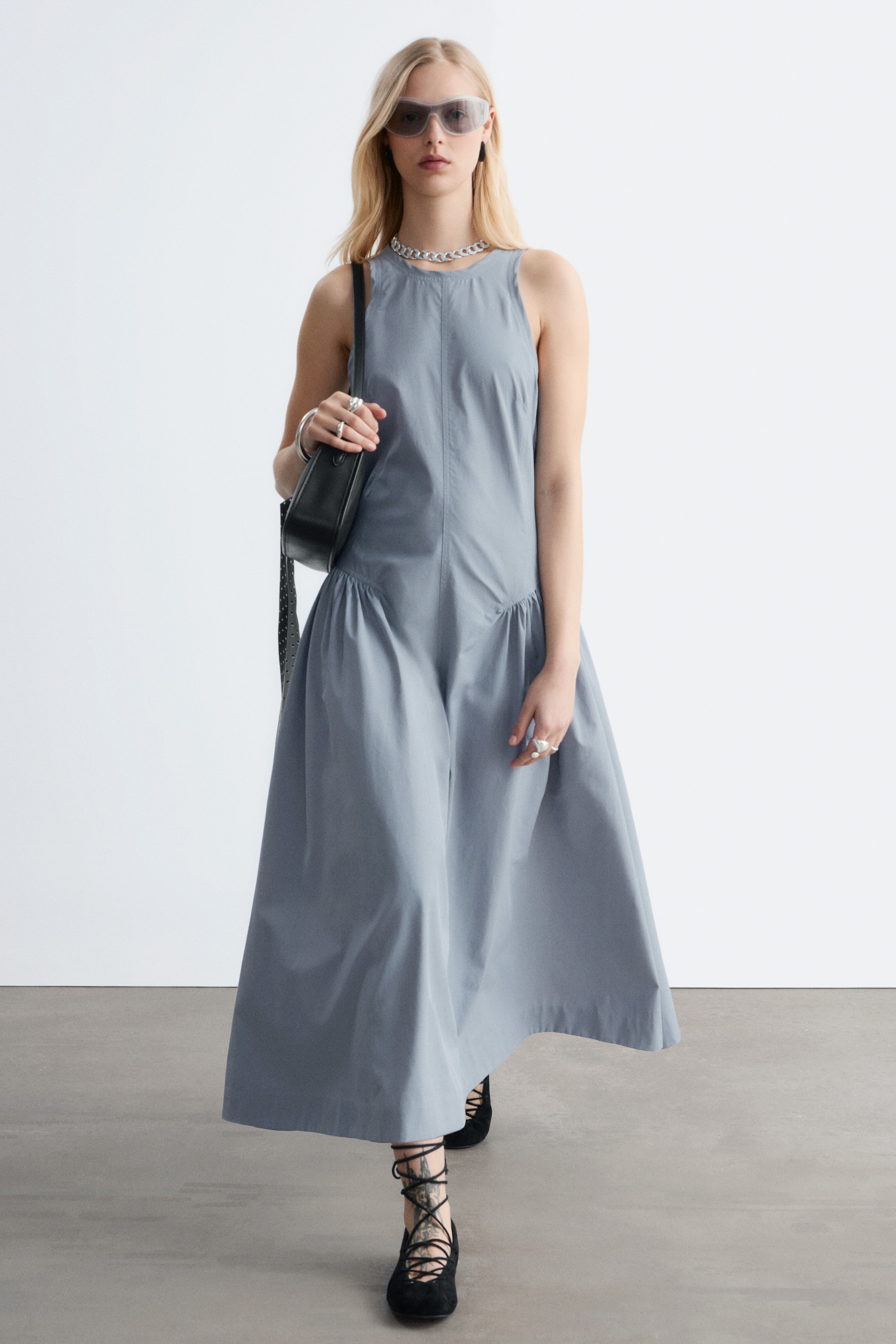 Se større billede: En gråblå ærmeløs kjole med rund halsudskæring og en sænket talje med en samlet, fyldig midi-nederdel. Den præsenteres forfra og viser sin flydende silhuet.