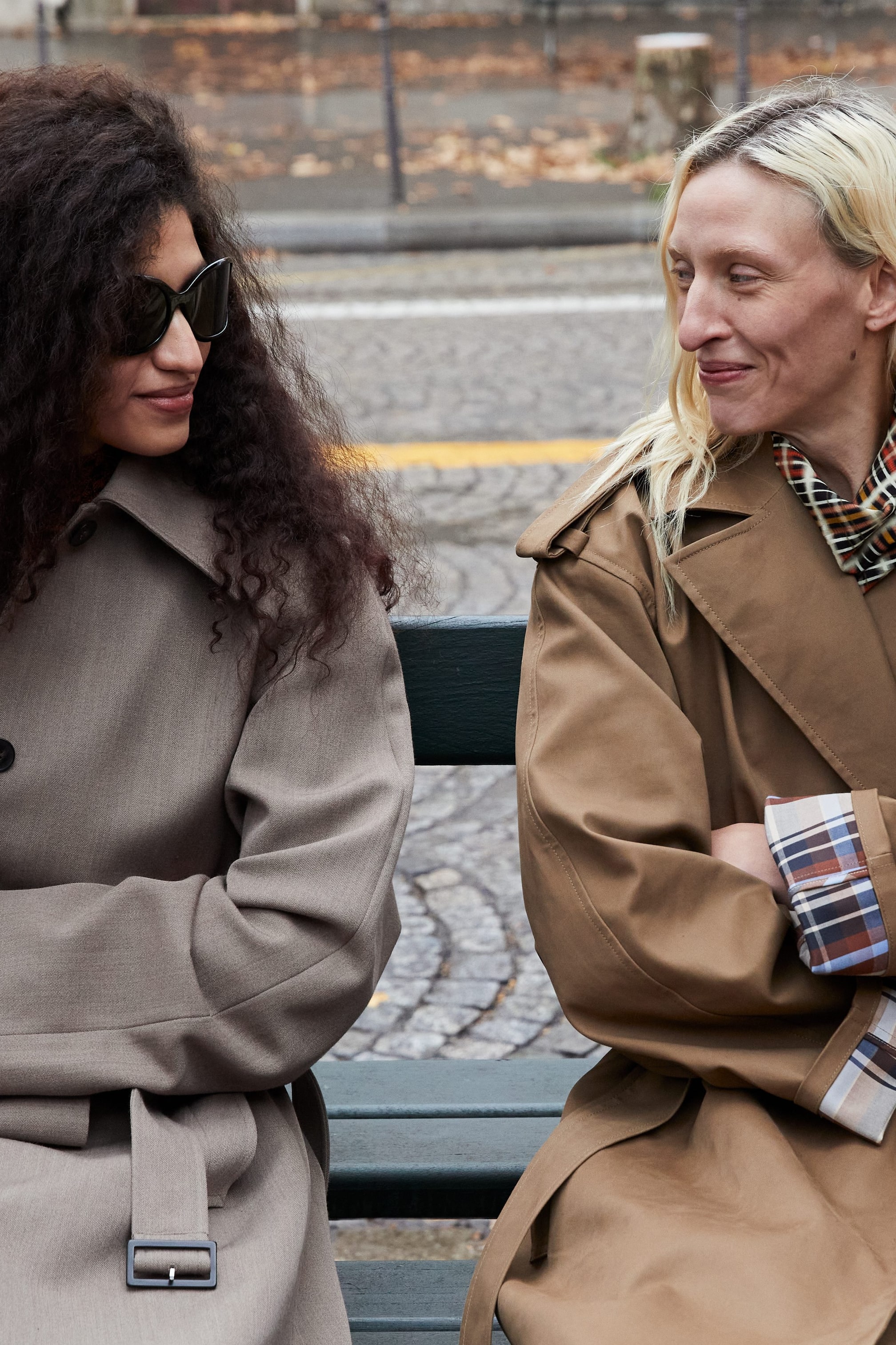 Grösseres Bild ansehen: Ein hellgrauer Trenchcoat mit einem einreihigen Design, sichtbaren Knöpfen und einem Gürtel mit rechteckiger Schnalle, getragen von einer Person, die nach rechts schaut. Ein beiger Trenchcoat wird ebenfalls präsentiert, mit Schulterklappen und hochgekrempelten Manschetten, die ein kariertes Futter zeigen, gestylt mit einem gemusterten Schal um den Hals, getragen von einer Person, die nach links schaut.