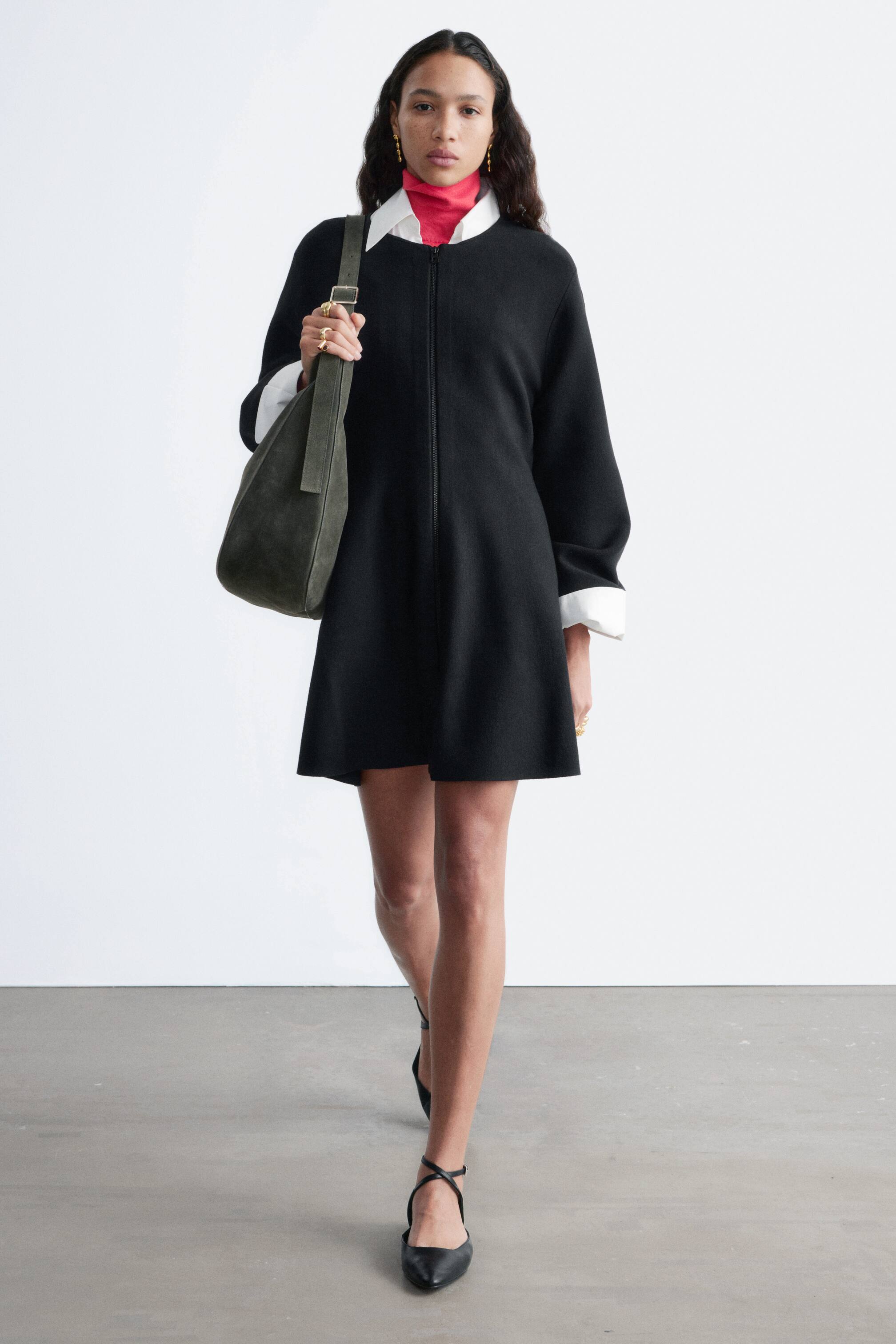 Ver imagen más grande: Un vestido negro de línea A en versión mini, con una cremallera frontal de largo completo. Está superpuesto sobre una camisa blanca con cuello y un jersey de cuello alto rosa, con el cuello y los puños blancos extendiéndose visiblemente desde el escote y las mangas del vestido.