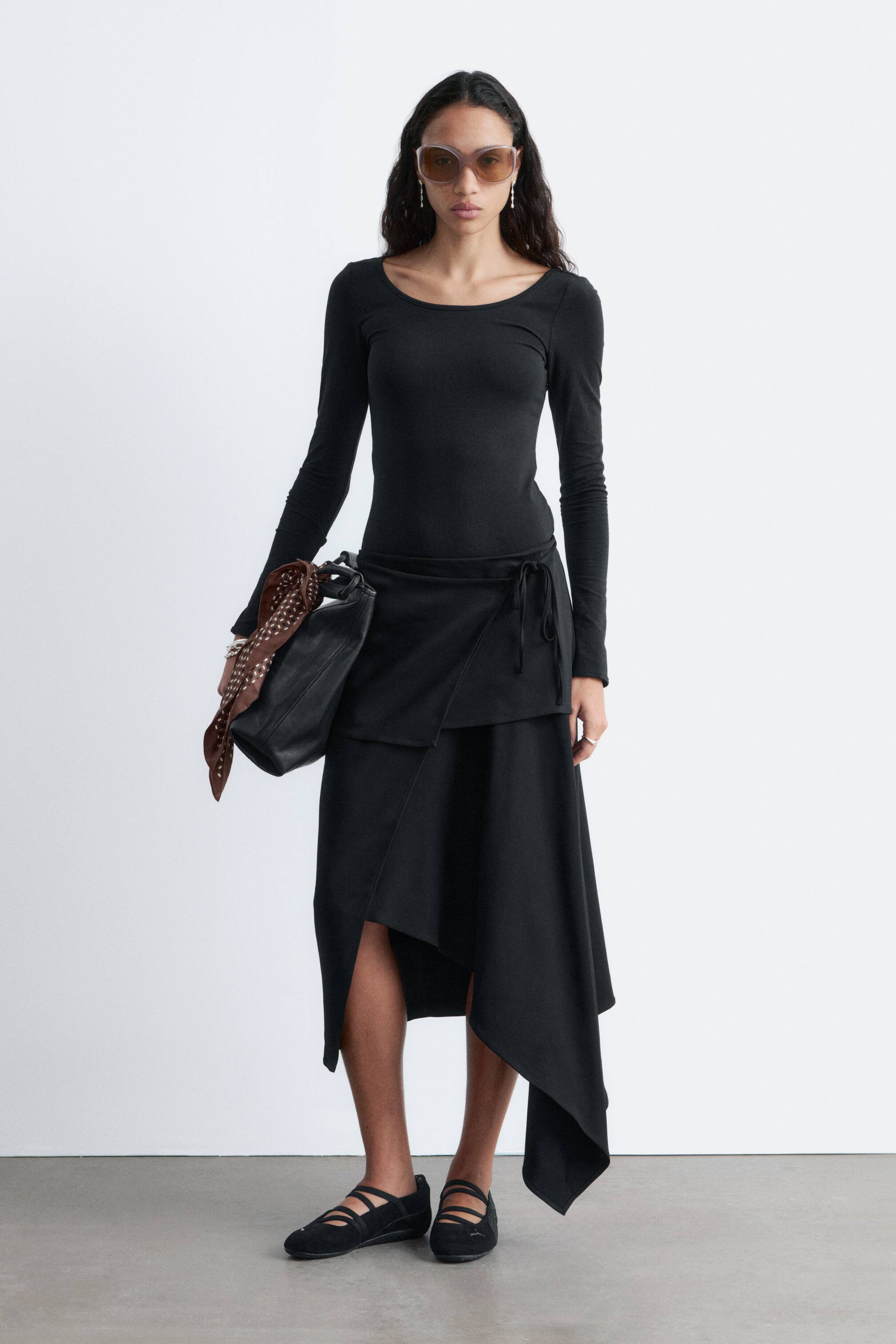 Vezi imaginea mai mare: O fustă neagră petrecută, cu tiv asimetric și cordon la șoldul stâng. Fusta se drapează lejer, creând un panou frontal suprapus și o deschidere pe piciorul stâng.