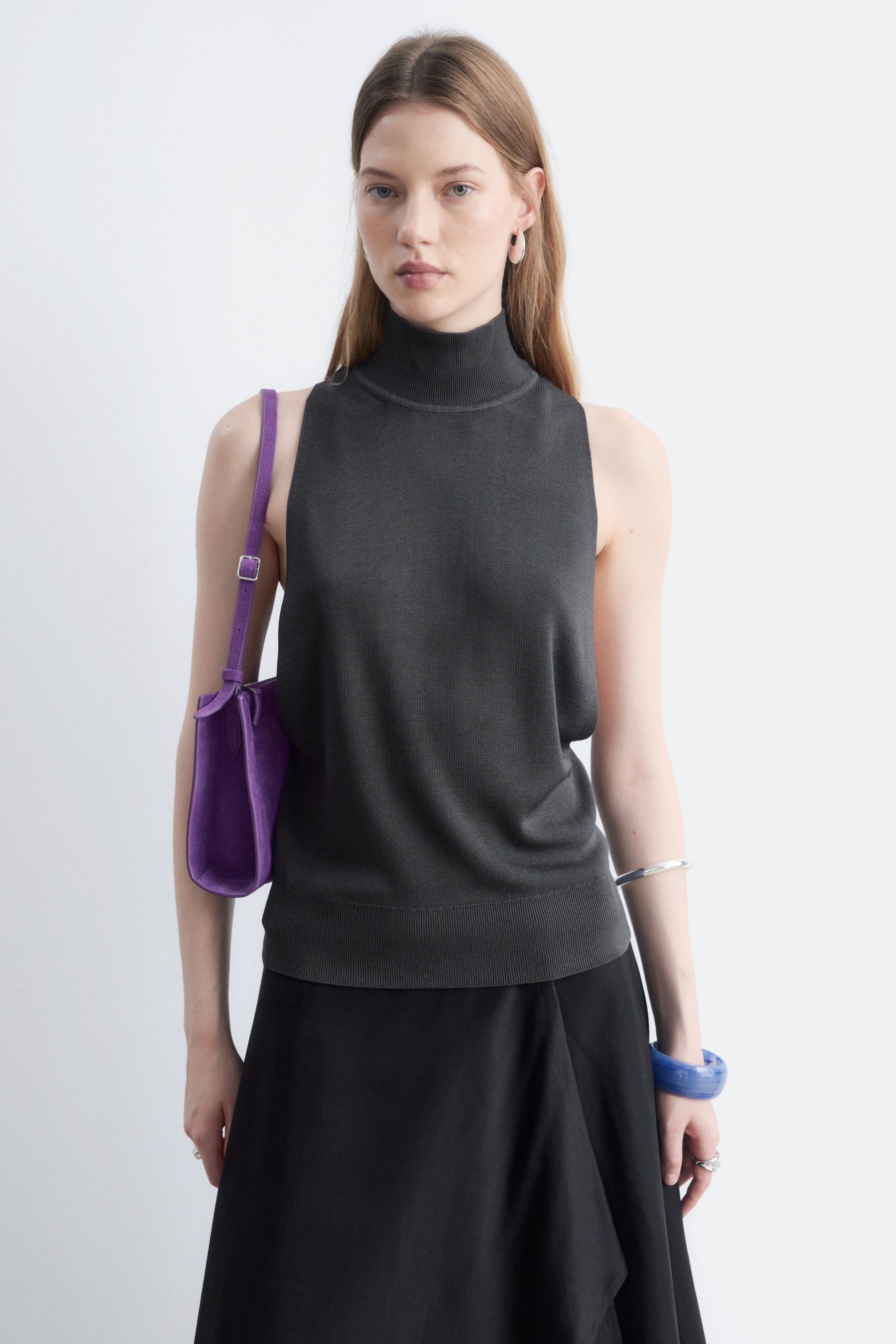 View larger image: A dark grey knitted sleeveless roll-neck top with a ribbed hem is being worn by the model, layered over a black skirt. The model is also carrying a purple shoulder bag.