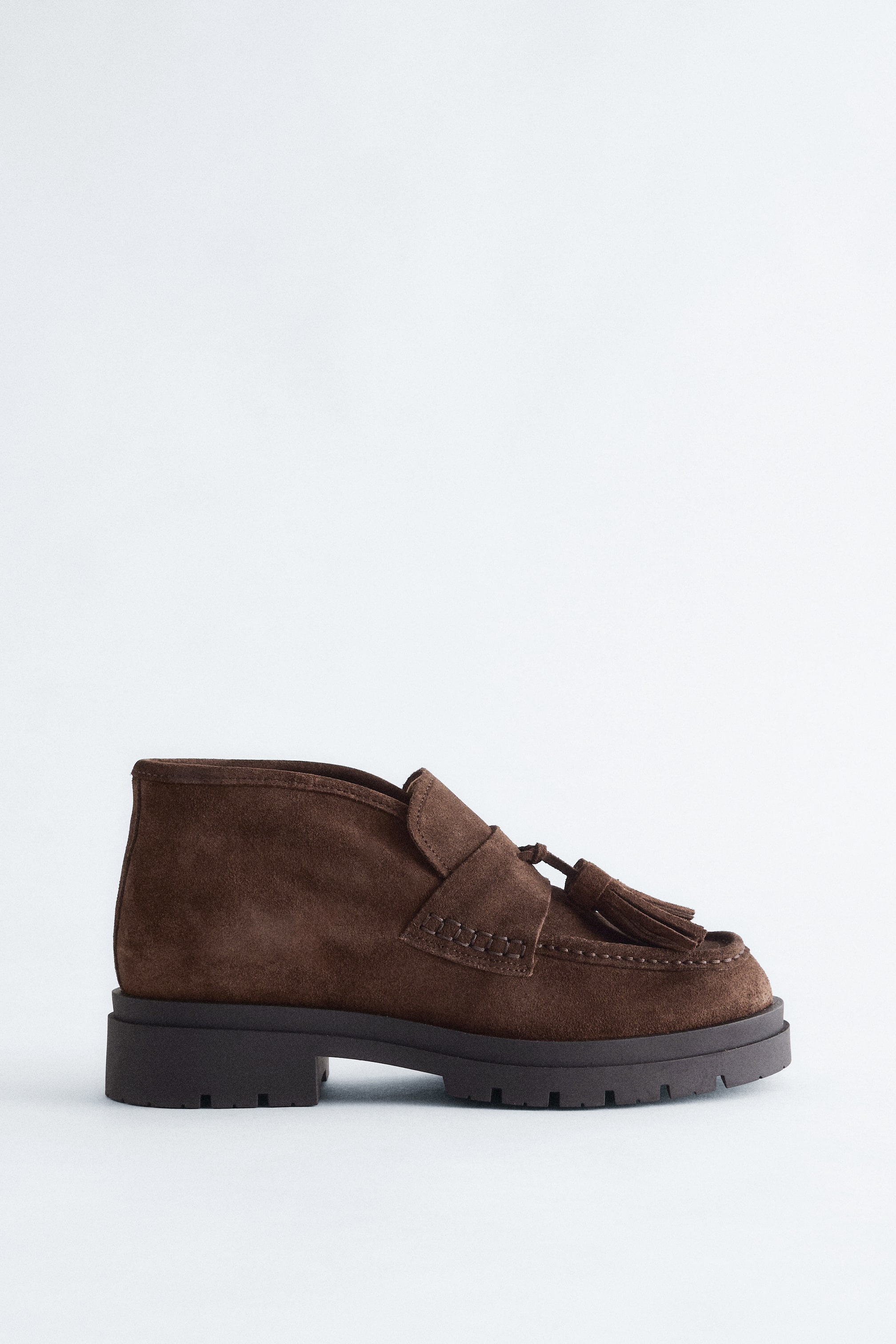 Ver imagen más grande: Un zapato de estilo mocasín de ante marrón está orientado hacia la izquierda, mostrando un eje a la altura del tobillo, una punta redondeada con una costura de mocasín y borlas en la parte superior. Está terminado con una suela gruesa y dentada.
