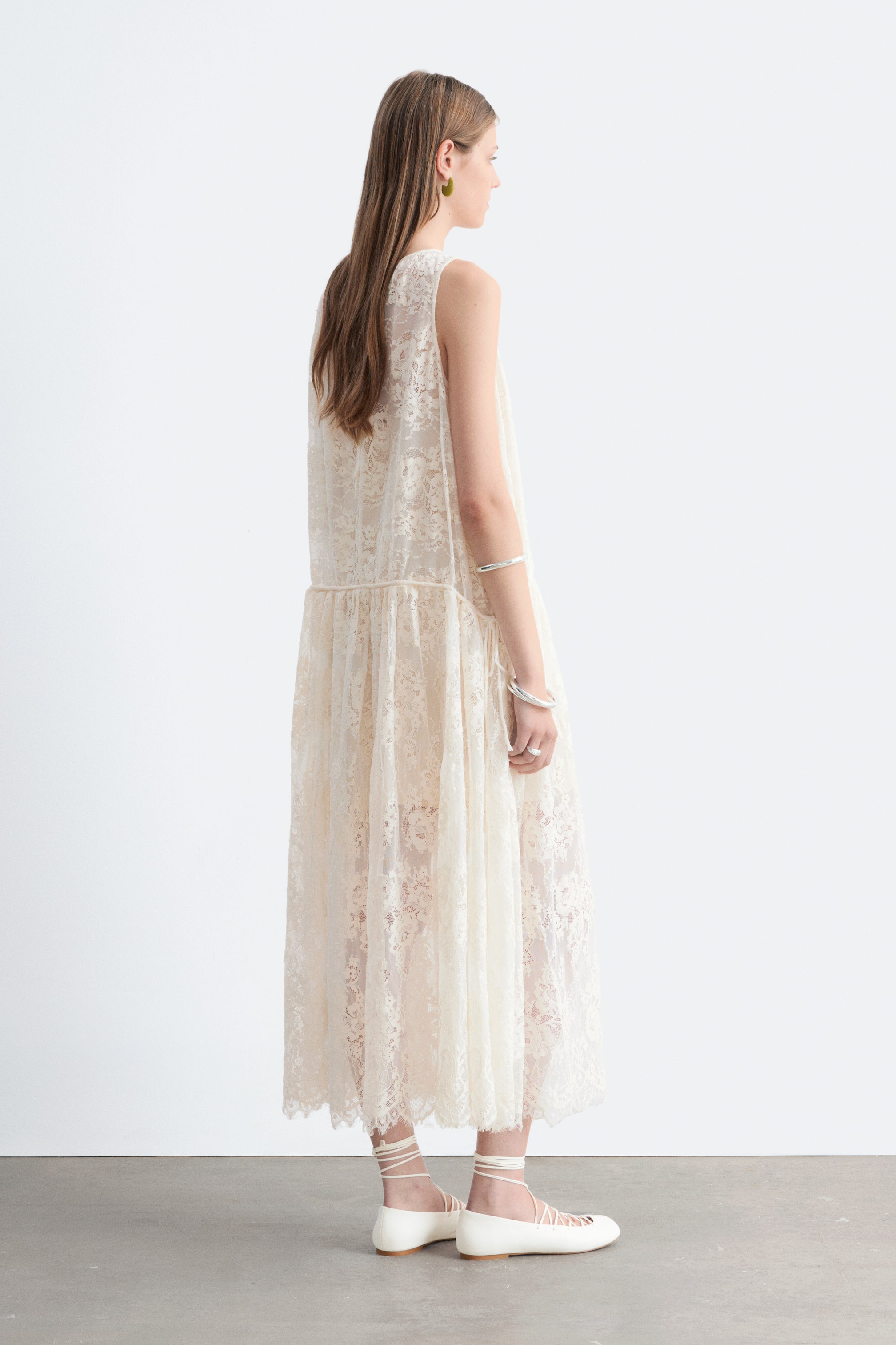 Größeres Bild ansehen: Ein cremefarbenes Spitzen-Maxikleid wird von dem Model getragen. Das ärmellose Kleid zeigt ein florales Spitzenmuster und eine geraffte, tief sitzende Taille, die bis zu den Knöcheln reicht, und wird durch weiße Ballerinas mit Schnürung komplettiert.