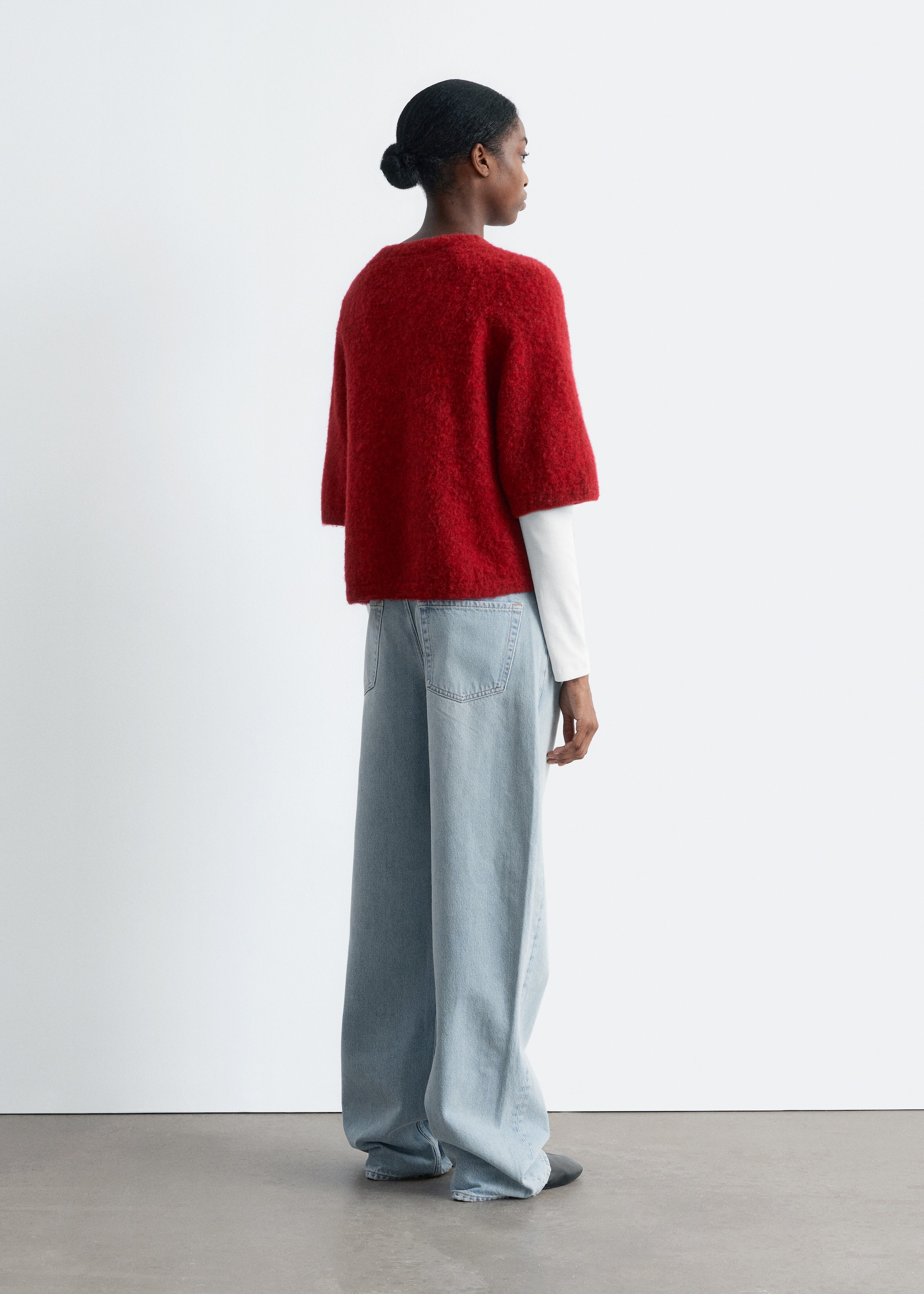 Grösseres Bild ansehen: Ein roter, flauschiger Strickpullover mit weiten, ellbogenlangen Ärmeln und einem runden Halsausschnitt wird getragen, von hinten betrachtet. Er ist über einem weißen langärmligen Hemd geschichtet und wird mit hellblauen, weit geschnittenen Jeans kombiniert, die sichtbare aufgesetzte Gesäßtaschen aufweisen.