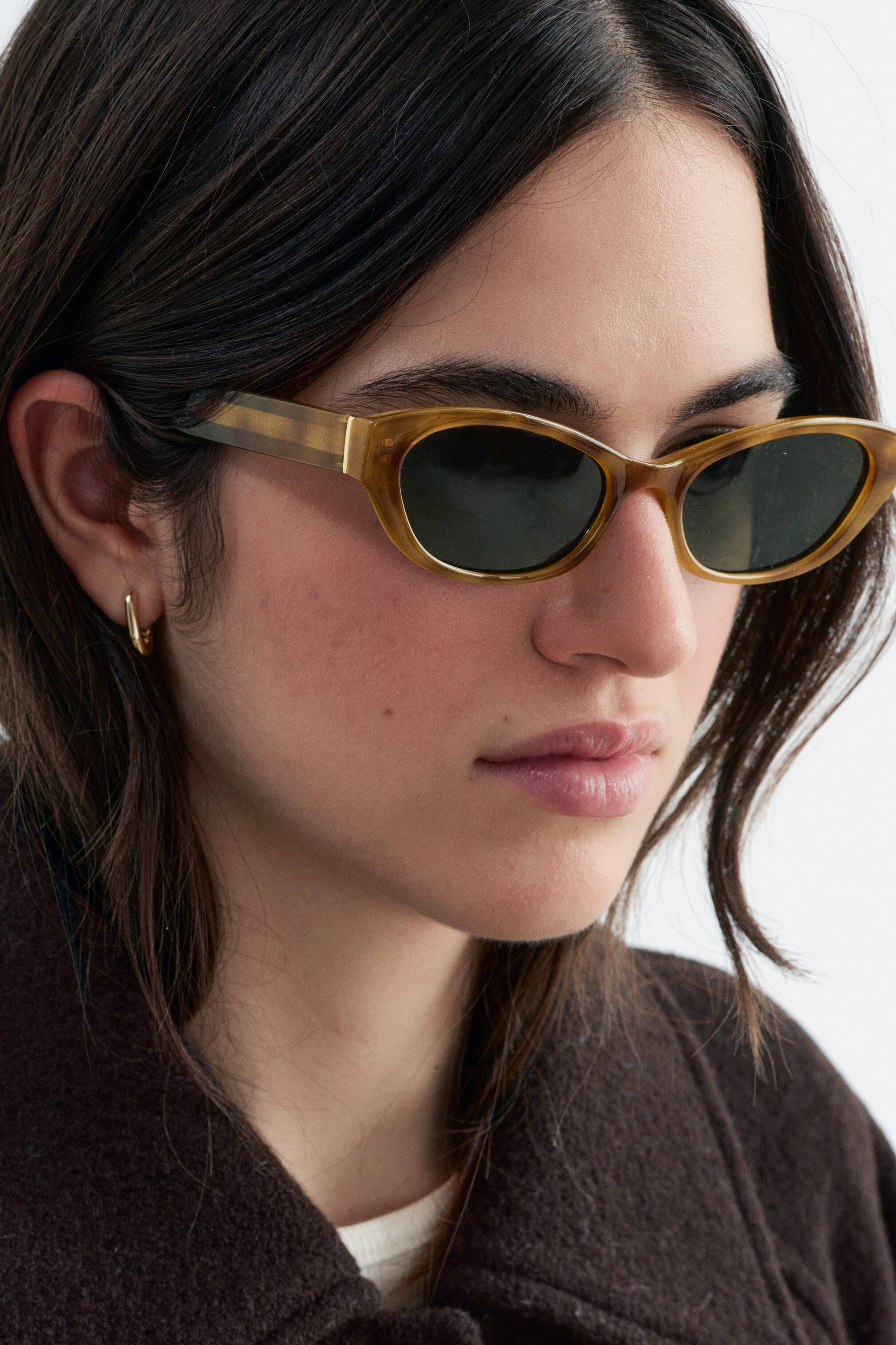 Ver imagem maior: Óculos de sol estreitos e ovais, com armações translúcidas de tom mel e lentes escuras, estão a ser usados pela modelo. Os óculos de sol combinam com um pequeno brinco de argola dourado e um casaco castanho-escuro.