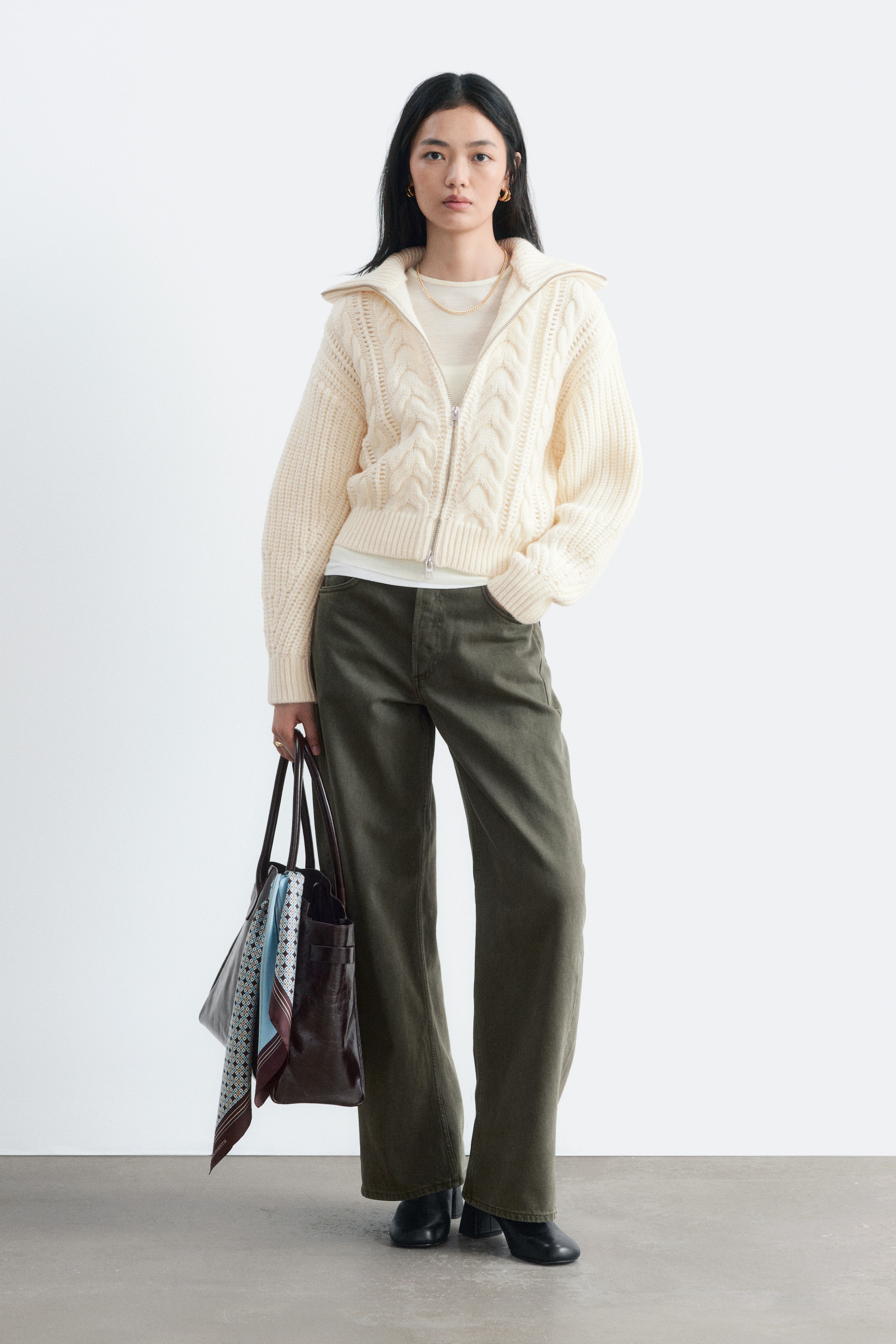 Visualizza immagine più grande: Jeans a gamba barrel in verde oliva, dal fit oversize, indossati dalla modella, che è in piedi con una mano appoggiata sulla tasca anteriore. I jeans a vita media sono abbinati a un maglione corto color crema lavorato a trecce con zip, un top bianco e stivali neri.