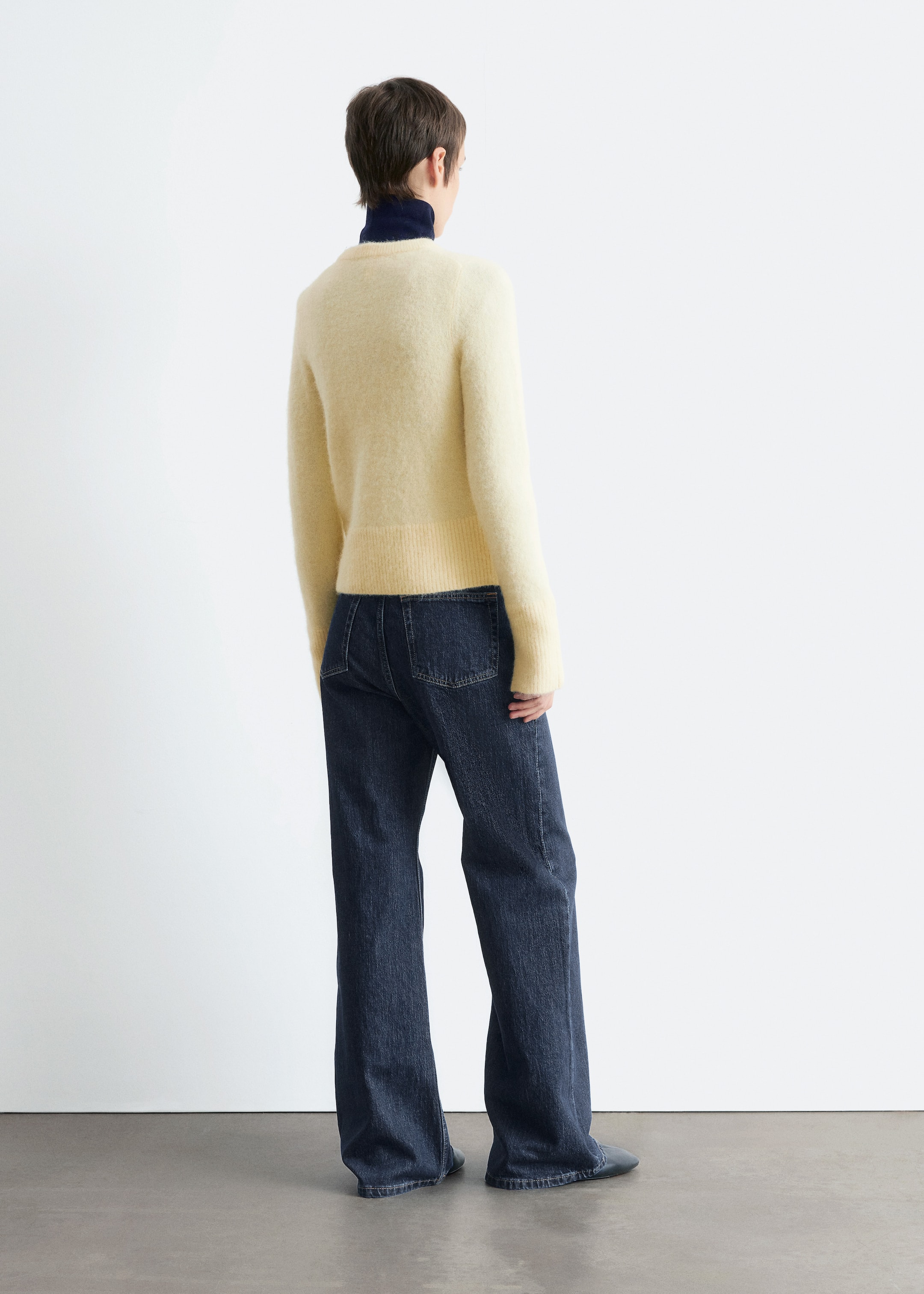 Ingrandisci l'immagine: Da dietro, un maglione girocollo giallo chiaro e peloso è indossato sopra un dolcevita blu scuro, con l'orlo a coste che si trova in vita. Jeans blu scuro a gamba larga completano il maglione, con due tasche posteriori applicate.