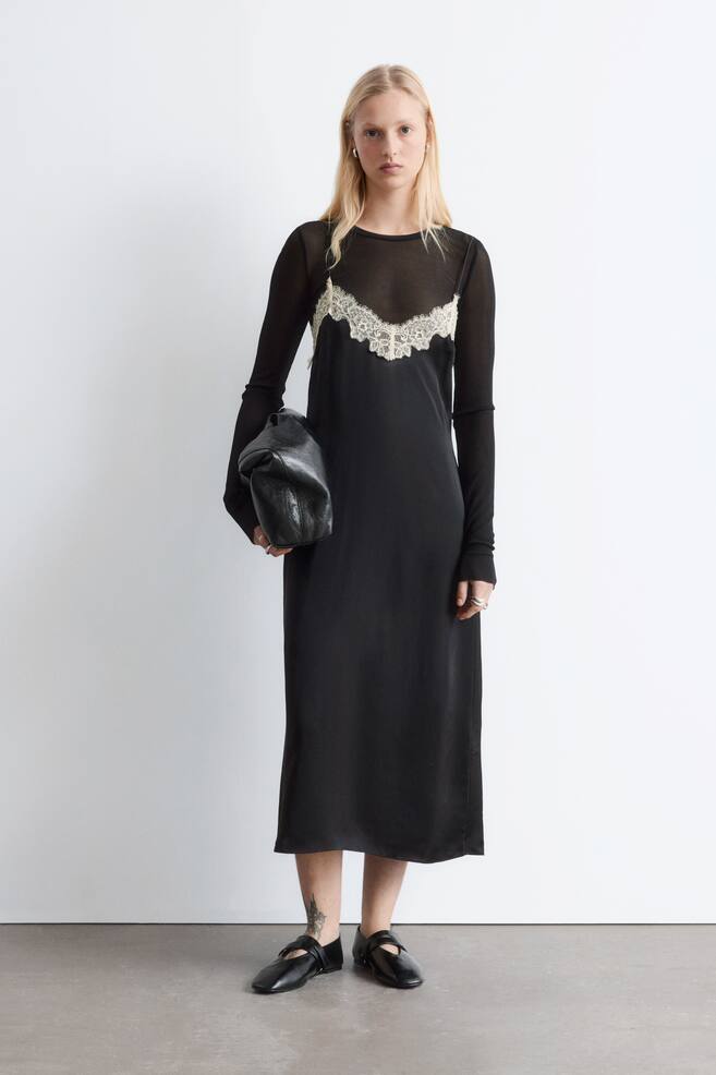 La robe noire midi à bretelles est présentée de face, superposée à un haut noir transparent à manches longues. La robe à bretelles arbore un décolleté en V avec des détails en dentelle blanc cassé et de fines bretelles, et tombe à mi-mollet. - 1
