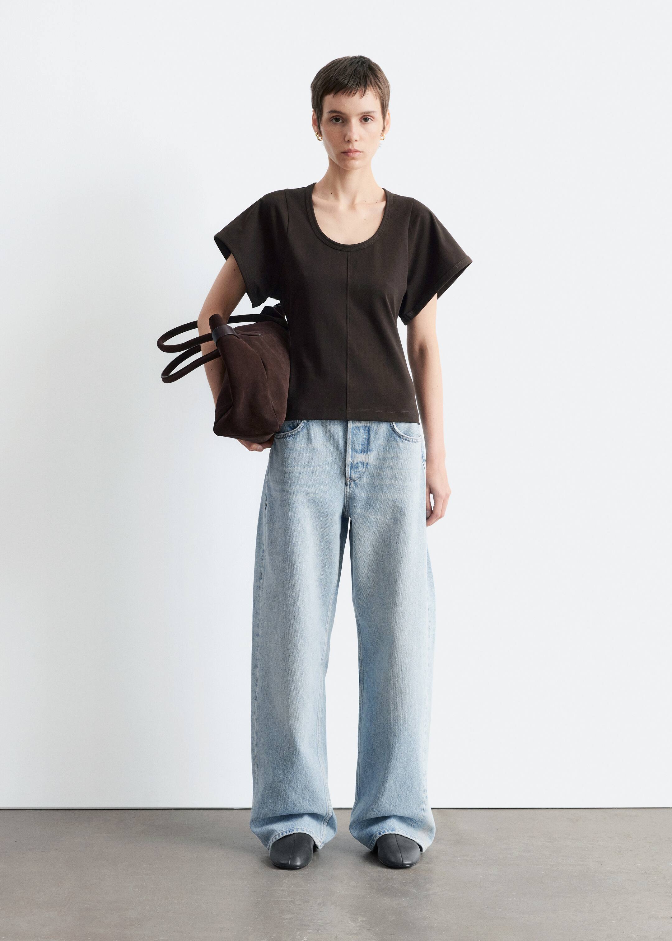 Agrandir l'image: Un haut marron foncé avec des manches courtes étendues, un col rond et une couture verticale centrale est porté par un mannequin, associé à un jean large à taille haute et délavé clair avec une braguette à boutons visible. Un sac texturé marron foncé avec deux poignées supérieures est tenu, et des chaussures à bout fermé foncées sont visibles.