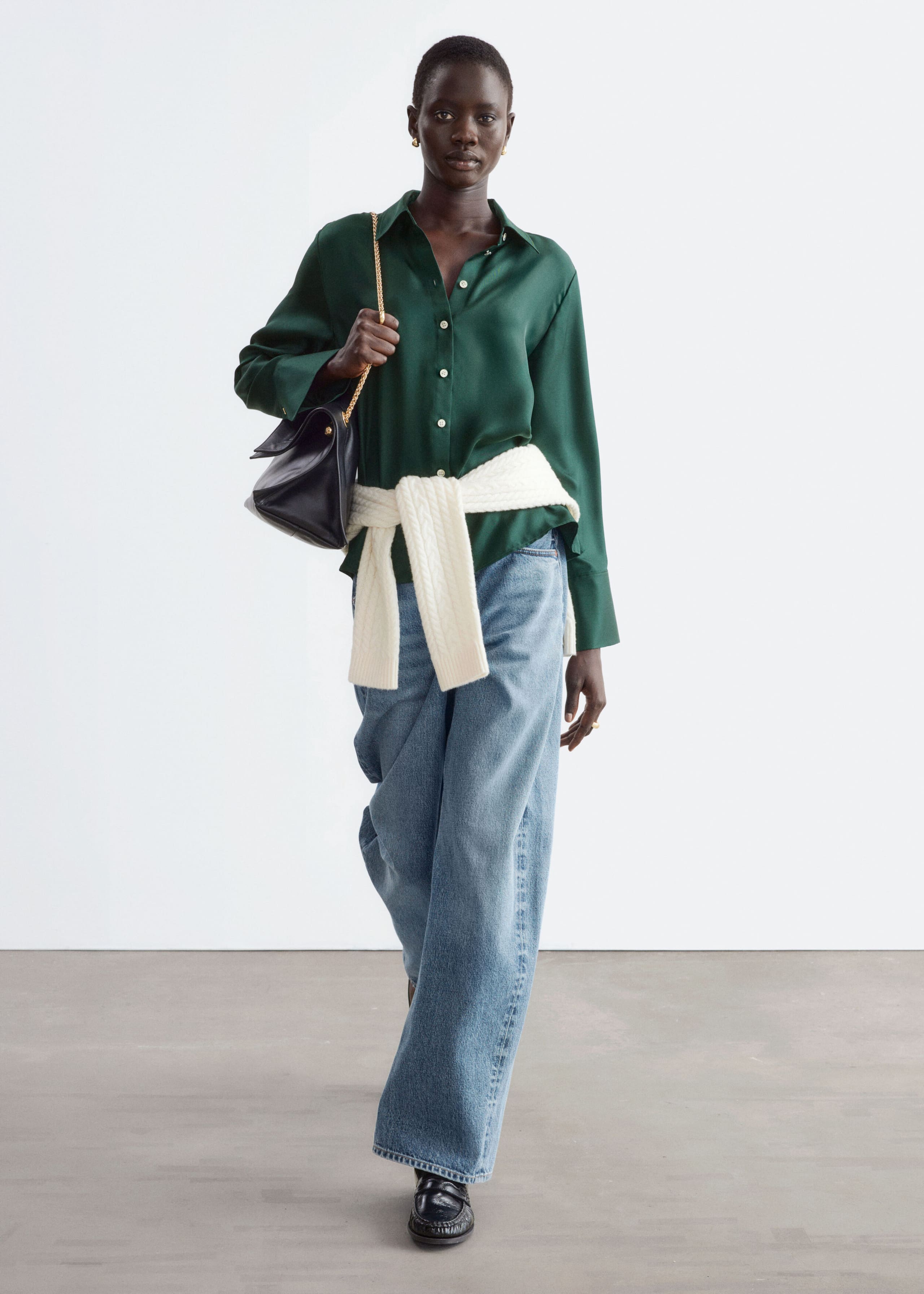 A dark green silk shirt with a lustrous finish, classic collar, button-down front, and wide cuffs is being worn by the model. The shirt is styled with wide-leg blue jeans, a cream-coloured jumper tied around the waist, and a black leather shoulder bag that the model is holding.