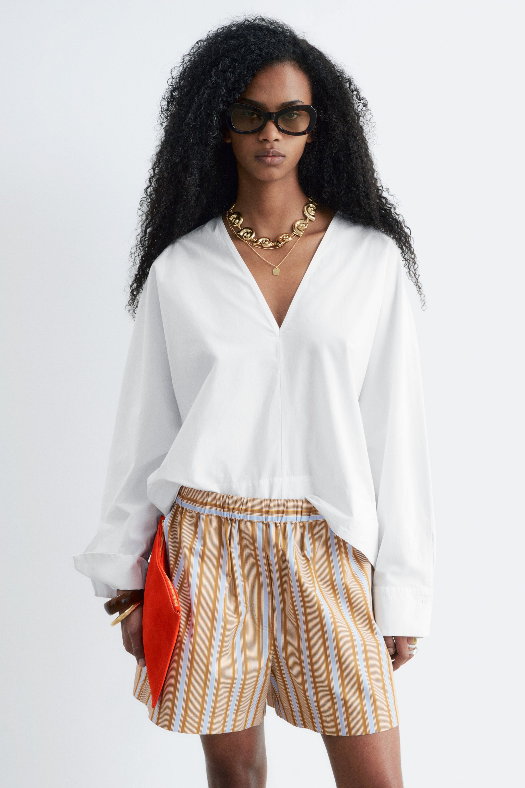 Grotere afbeelding bekijken: Een wit shirt met lange mouwen wordt gepresenteerd, met een diepe V-hals, verlaagde schouders en brede manchetten. Het wordt gedragen over gestreepte shorts, die verticale strepen in beige, lichtblauw en bruin tonen met een elastische tailleband.