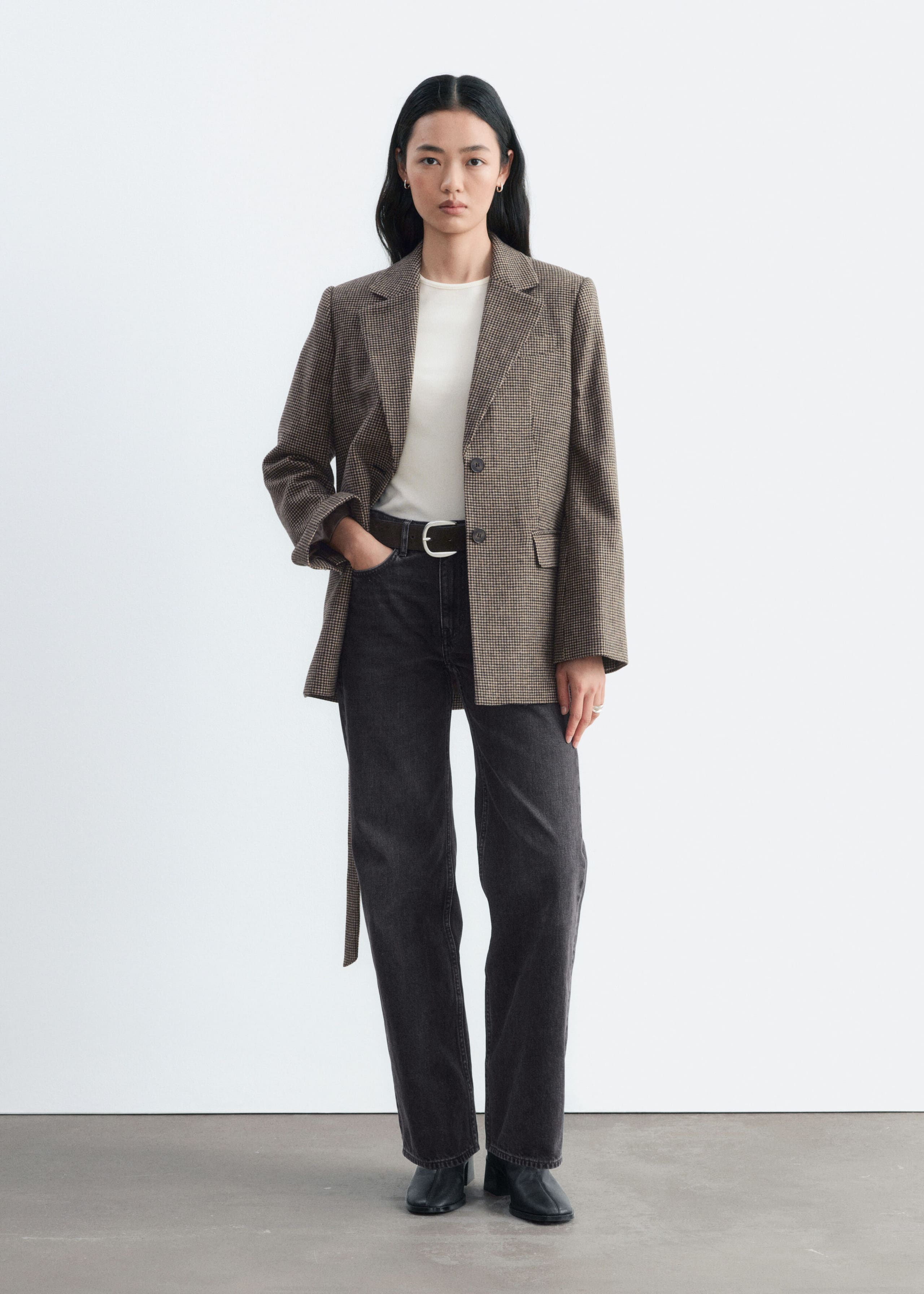 A brown houndstooth wool-blend blazer featuring notch lapels, flap pockets and a matching tie belt is worn open by the model. The blazer is layered over a cream-coloured top and paired with dark grey wide-leg jeans.