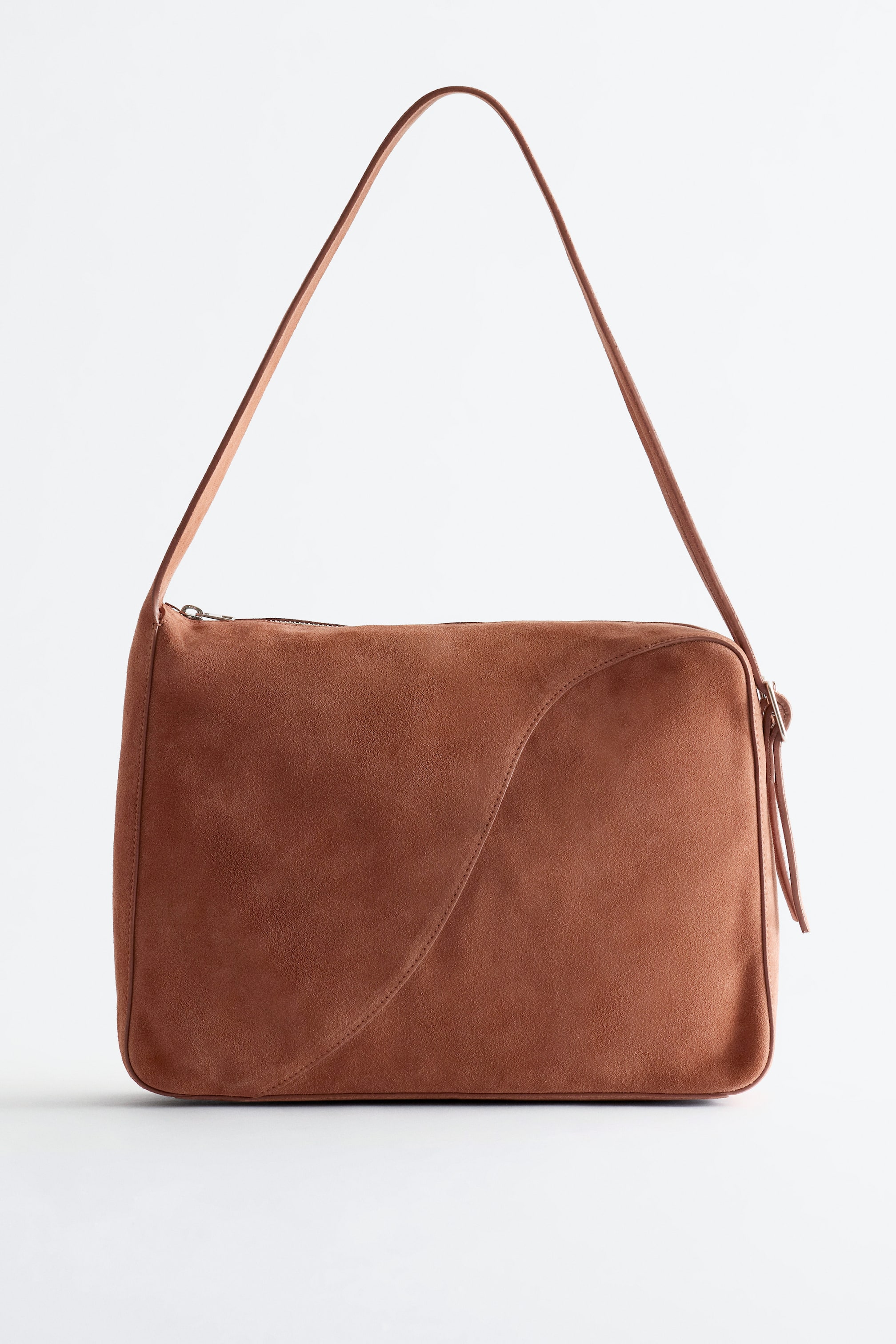 Προβολή μεγαλύτερης εικόνας: Μια καφέ σουέτ τσάντα tote παρουσιάζεται με πρόσοψη, διαθέτοντας ένα δομημένο, ορθογώνιο σώμα με λεία υφή και μια έντονη καμπύλη ραφή στο μπροστινό πάνελ της. Ένας λεπτός ιμάντας ώμου καμπυλώνεται πάνω από την τσάντα, συνδέοντας με μικρούς μεταλλικούς κρίκους στις επάνω γωνίες, και ένα κλείσιμο με φερμουάρ είναι ορατό κατά μήκος της επάνω άκρης.