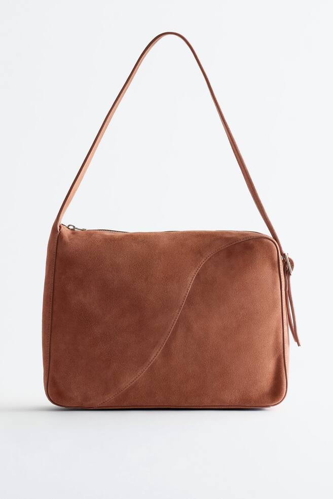 Un sac cabas en daim marron est présenté de face, doté d'un corps rectangulaire structuré avec une texture lisse et une couture incurvée proéminente sur son panneau avant. Une fine bandoulière s'arque au-dessus du sac, attachée à de petits anneaux métalliques aux coins supérieurs, et une fermeture éclair est visible le long du bord supérieur. - 1