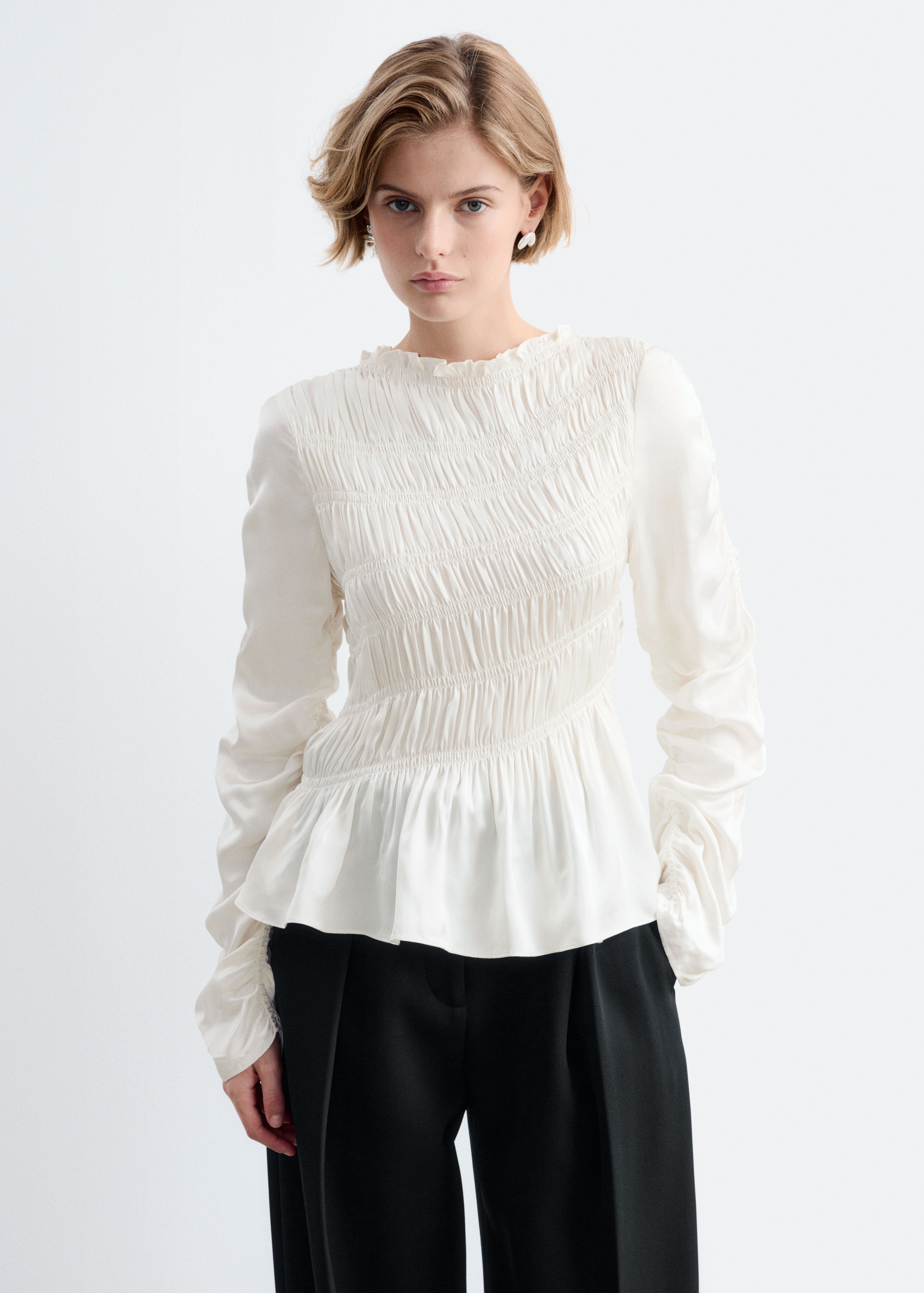 O bluzÄ din satin, de culoarea cremei, cu detalii smock, este orientatÄ spre faÈÄ, evidenÈiind un corsaj complet smock, cu guler Ã®nalt cu volane Èi tiv peplum evazat. MÃ¢necile lungi prezintÄ detalii cu fronseuri de-a lungul cusÄturilor exterioare.