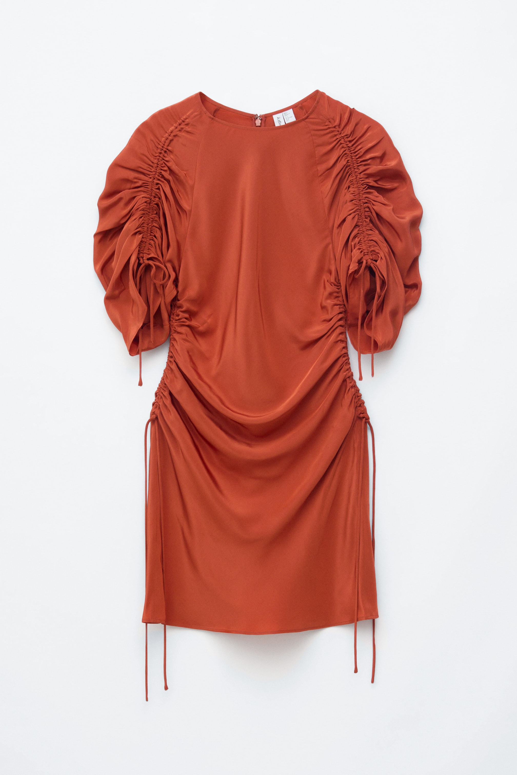 Agrandir l'image: Vue de FACE d'une robe courte couleur rouille présentant une silhouette fluide et drapée en tissu léger. Elle est dotée de manches courtes volumineuses et froncées avec des liens à nouer sur l'extérieur du bras, ainsi que de fronces verticales le long des coutures latérales, des emmanchures à l'ourlet, contrôlées par des liens intégrés.