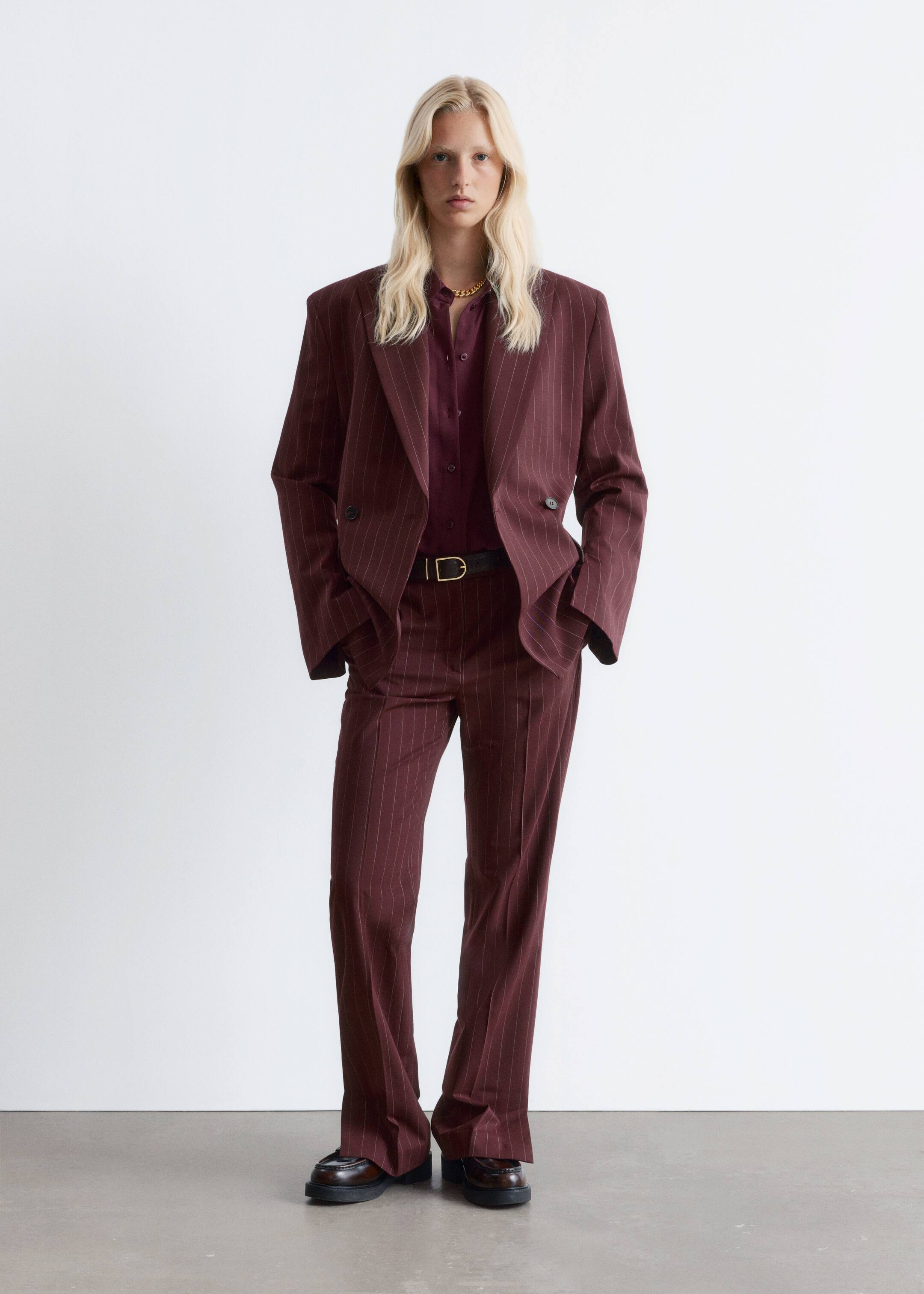Agrandir l'image: Un costume à rayures bordeaux est présenté, composé d'un blazer croisé surdimensionné avec des revers en pointe et des épaules rembourrées, et d'un pantalon large assorti. Une chemise bordeaux à manches longues est portée sous le blazer, cintrée par une ceinture noire, et des mocassins sombres sont visibles.