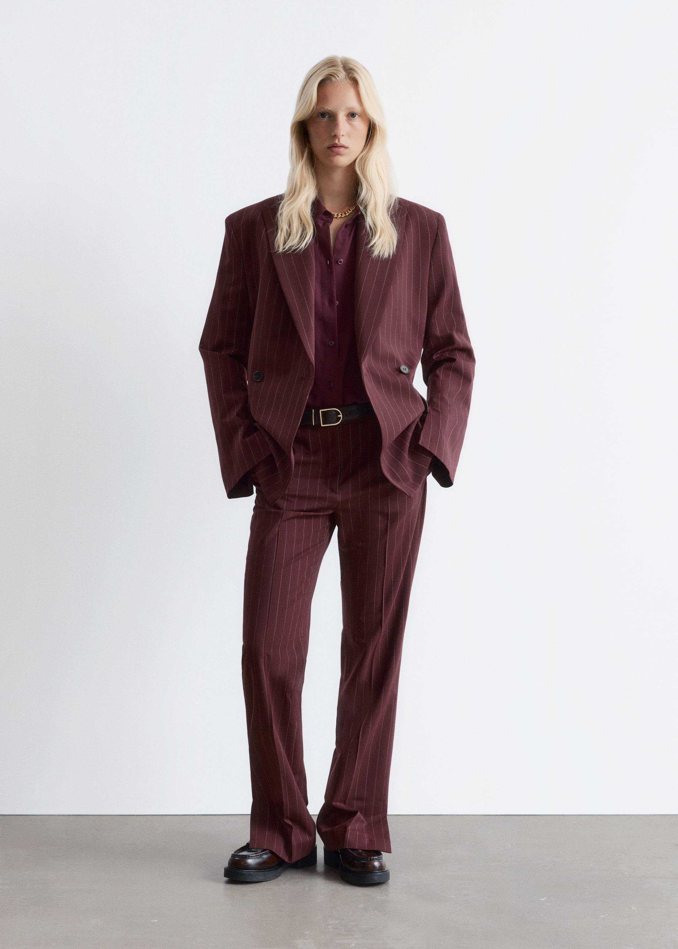 Grotere afbeelding bekijken: Een bordeauxrood krijtstreeppak wordt gepresenteerd, bestaande uit een oversized double-breasted blazer met inkeping revers en gewatteerde schouders, en bijpassende wijde broek. Onder de blazer wordt een bordeauxrood overhemd met lange mouwen gedragen, aangesnoerd door een zwarte riem, en donkere loafers zijn zichtbaar.