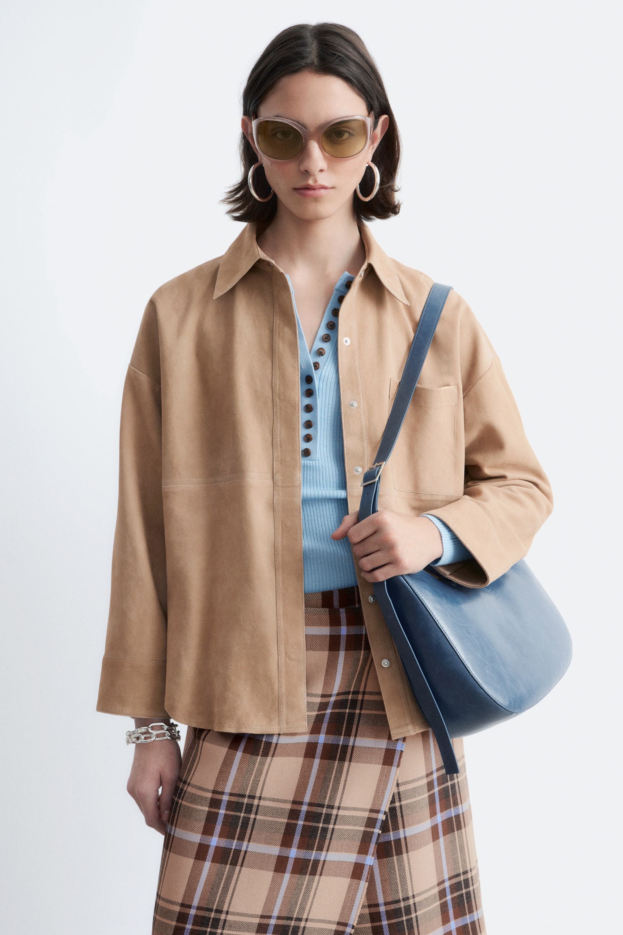 Ingrandisci l'immagine: Una borsa a tracolla blu a forma di mezzaluna, realizzata in un materiale liscio, è portata dalla modella. La modella tiene il corpo della borsa con una mano, mentre la tracolla regolabile è appoggiata sulla spalla.