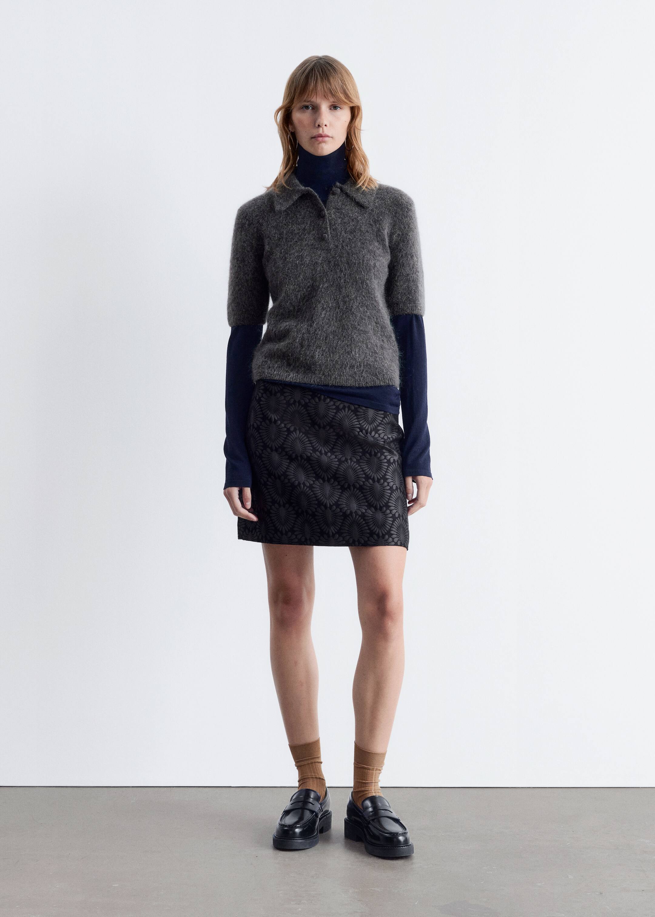 Größeres Bild ansehen: Ein grauer, kurzärmeliger, flauschiger Polopullover mit spitzem Kragen und einer kurzen Knopfleiste wird von vorne präsentiert. Dieser Pullover ist über einem dunkelblauen, langärmeligen Rollkragenpullover geschichtet und mit einem schwarzen Minirock gestylt, der ein durchgehendes geprägtes Fächer-Muster aufweist. Das Ensemble wird mit schwarzen, klobigen Loafern und hellbraunen, gerippten Socken vervollständigt.