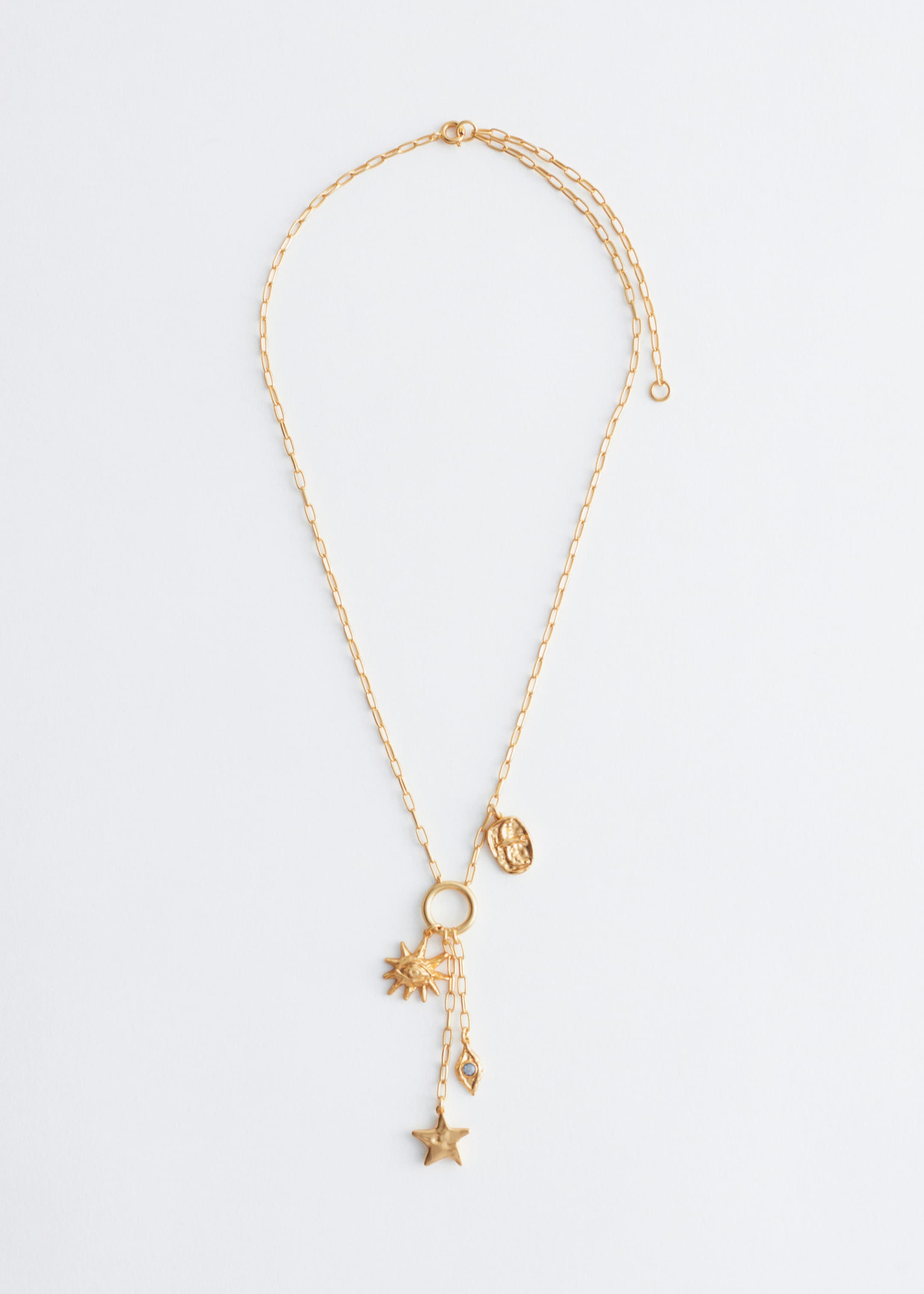 View larger image: A polished brass lariat-style necklace is lying flat, facing FRONT, displaying a paperclip chain with an oval charm attached to its upper section and a central ring below, from which textured figurative charms are suspending, including a sun with an eye, a star, and a teardrop. A spring ring clasp and an adjustable length chain are also visible at the top right.