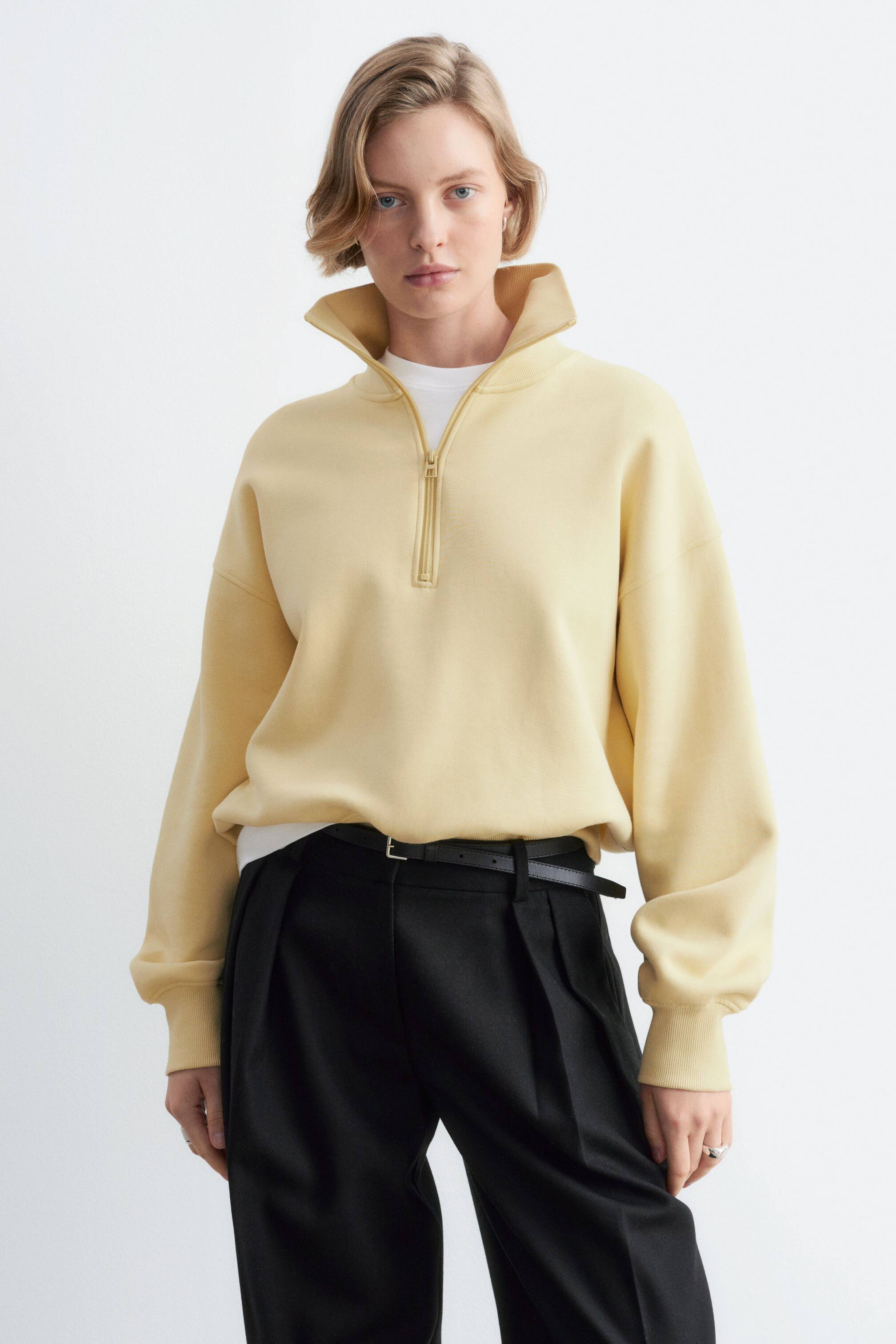 Visa större bild: En ljusgul sweatshirt i bomullsjersey med halv dragkedja och hög krage bärs av modellen. Sweatshirten i avslappnad passform har nedhasade axlar med ribbade muddar och är stylad i lager över en vit T-shirt och instoppad i svarta byxor.