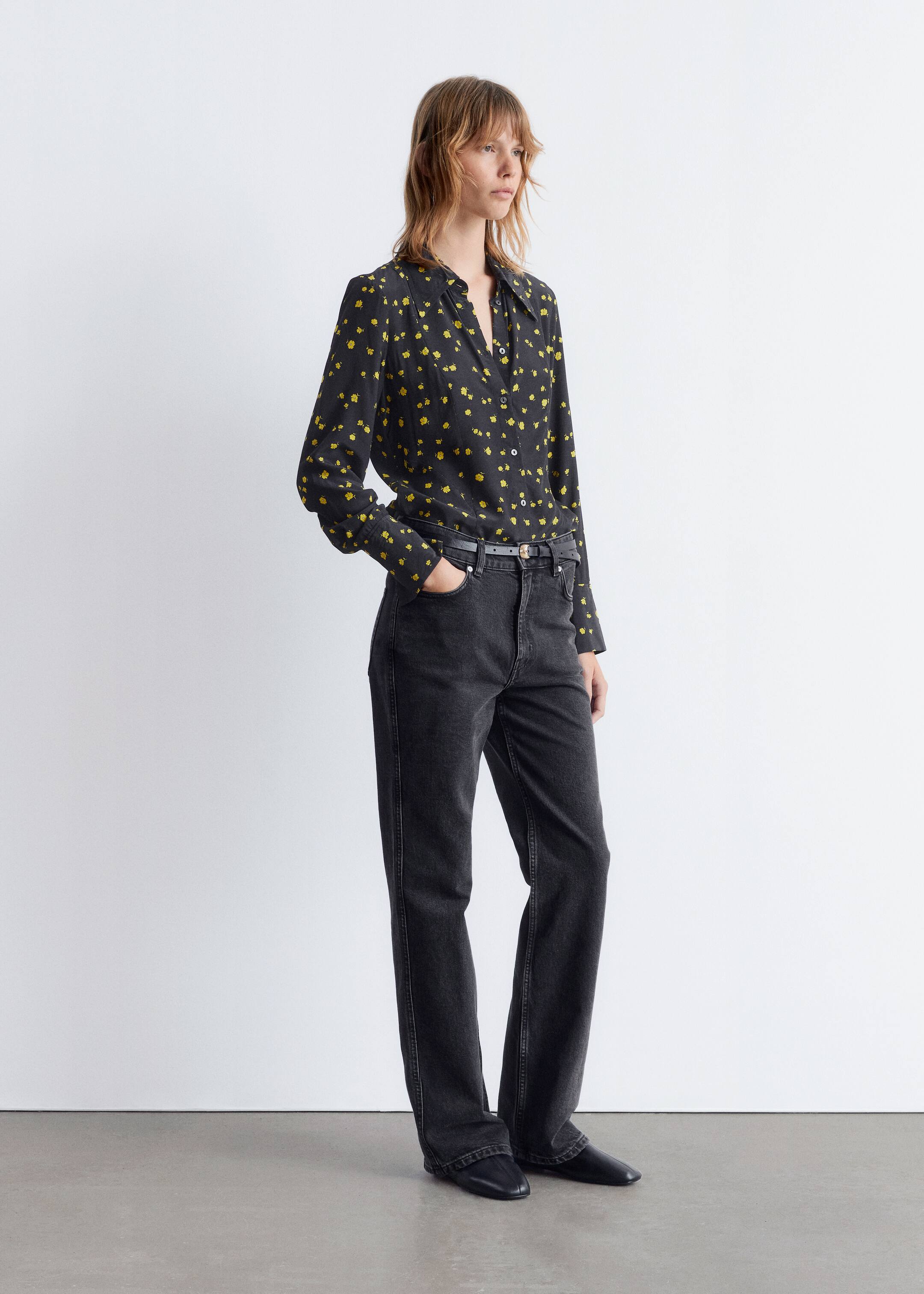 Ver imagem maior: Uma camisa preta de manga comprida com um padrão floral abstrato amarelo, gola pontiaguda e punhos com botões está a ser usada. A camisa está parcialmente enfiada em jeans pretos lavados, de cintura alta e perna reta, que são segurados por um cinto preto fino, com sapatos pretos de bico quadrado visíveis abaixo.