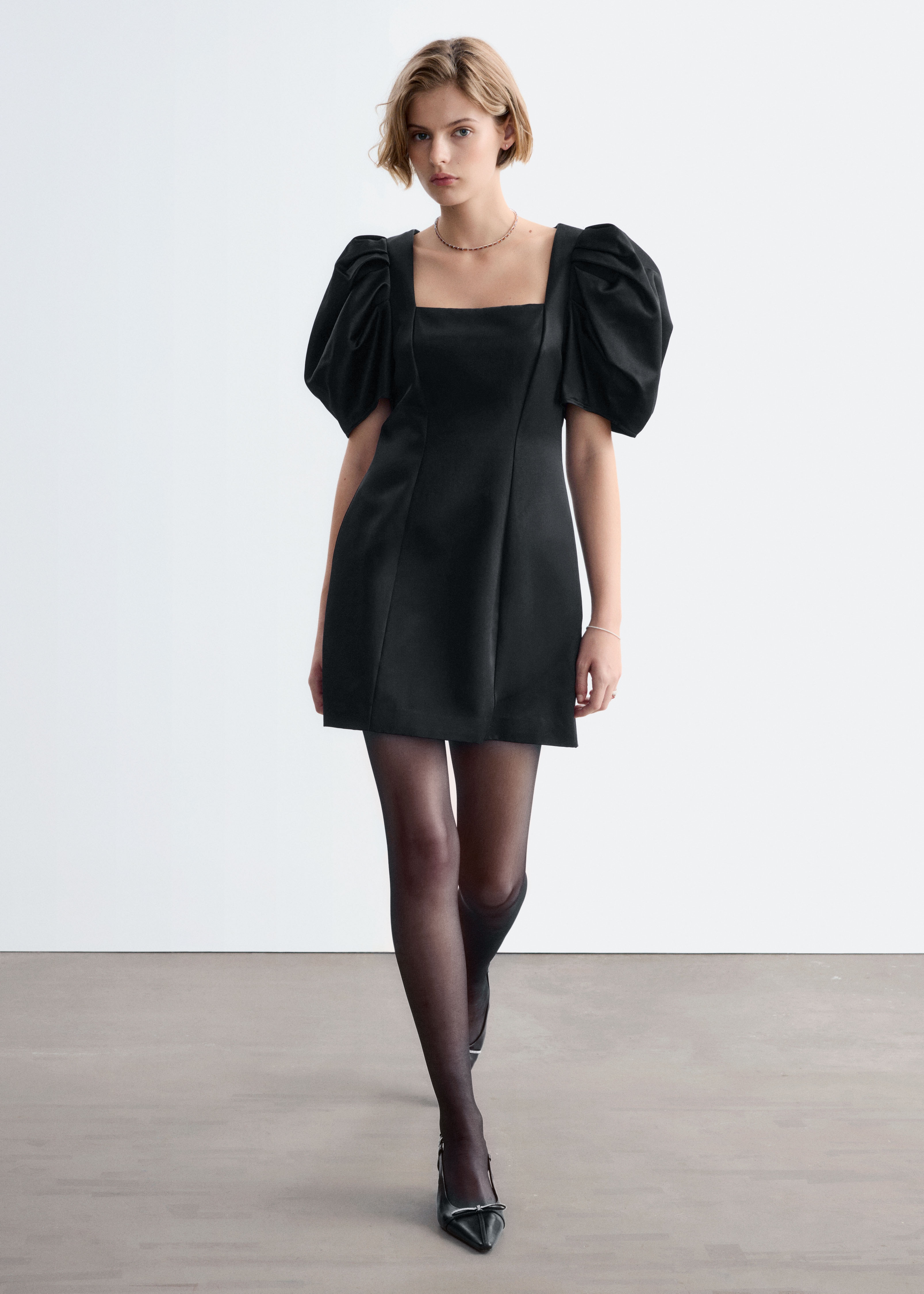 O rochie mini din satin negru este prezentatÄ din faÈÄ, avÃ¢nd un decolteu pÄtrat Èi mÃ¢neci scurte, bufante, pliate. Rochia afiÈeazÄ o siluetÄ structuratÄ, slim-fit, cu cusÄturi verticale care coboarÄ pe partea din faÈÄ.