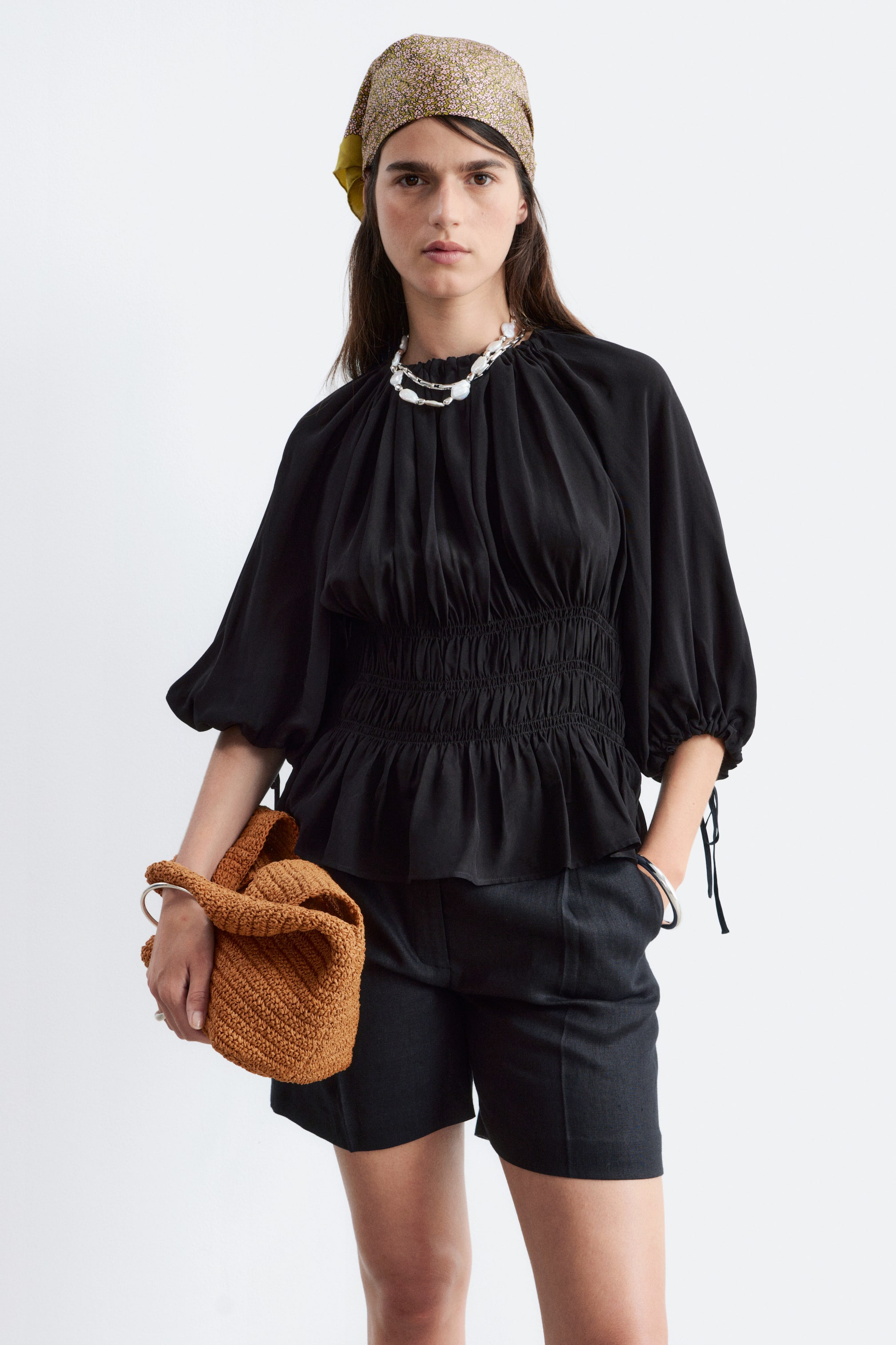 Größeres Bild ansehen: Eine schwarze Bluse mit einem gerafften hohen Ausschnitt, über dem eine silber-weiße Perlenkette liegt. Sie ist mit voluminösen Dreiviertelärmeln gestaltet, die mit Bindemanschetten gerafft sind, und ein breiter gesmokter Bund erzeugt einen Schößchensaum.