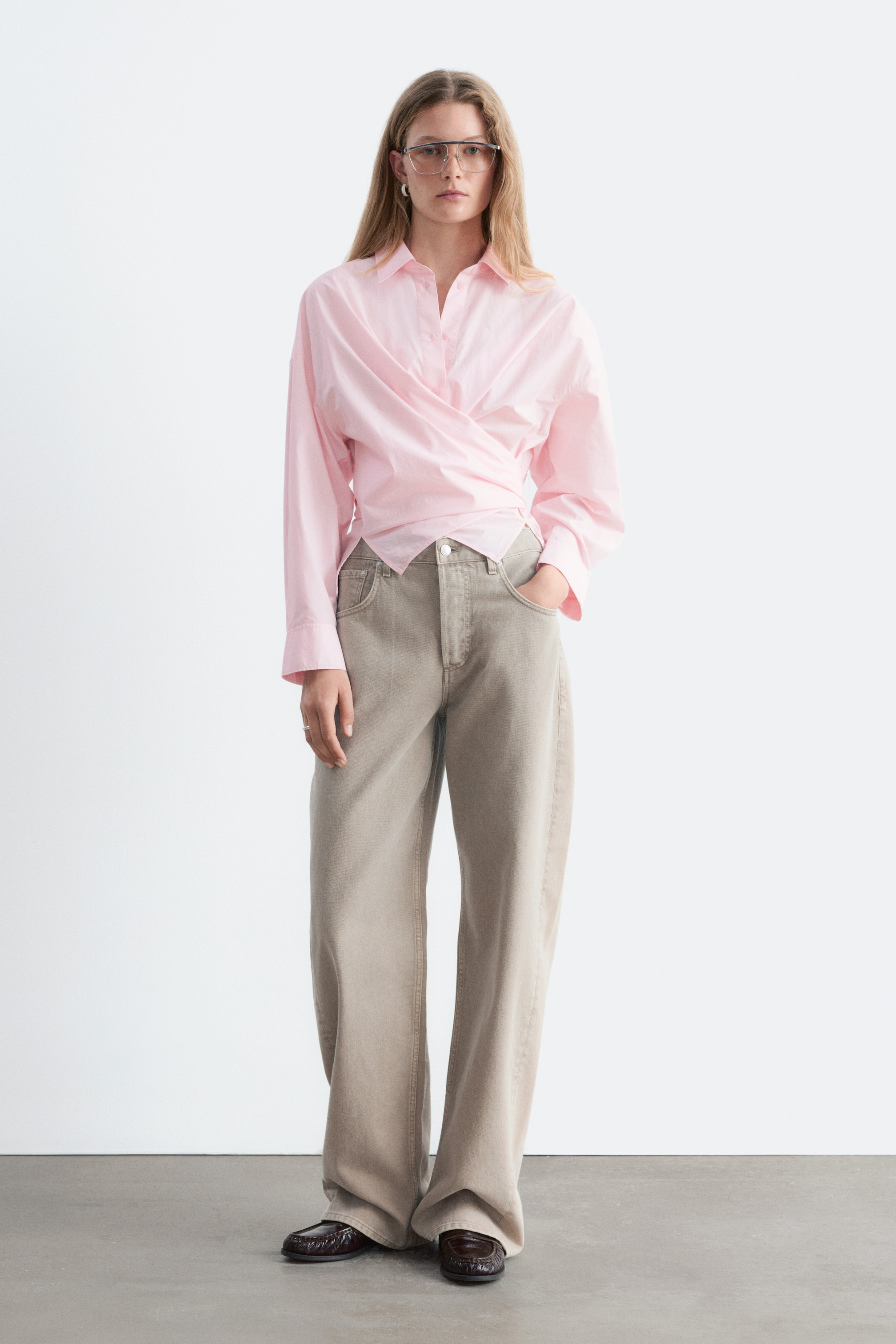 Agrandir l'image: La chemise en coton rose clair à nouer à la taille est présentée de face, avec un col pointu, des épaules tombantes et des manches longues à poignets boutonnés. Les pans avant de la chemise se croisent sur le buste et se nouent à la taille, créant une silhouette drapée et croisée qui tombe au-dessus d’un pantalon à jambes larges.