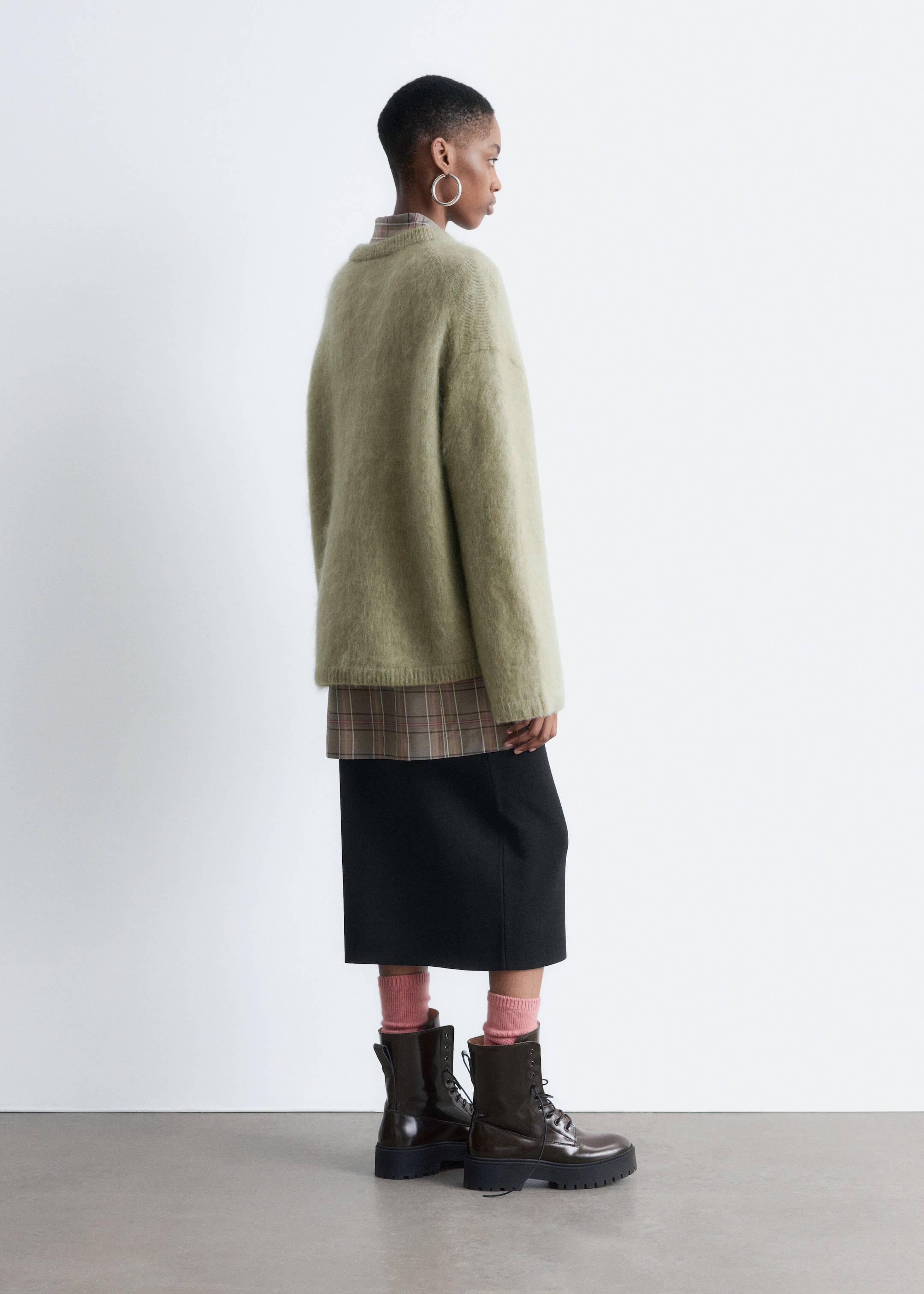 Visa större bild: En ljusgrön tröja i mohair- och ullblandning bärs av modellen. Den ledigt sittande tröjan är designad med ribbad hals, ärmslut och fåll, och den är lagrad över en rutig skjorta och en svart midikjol.