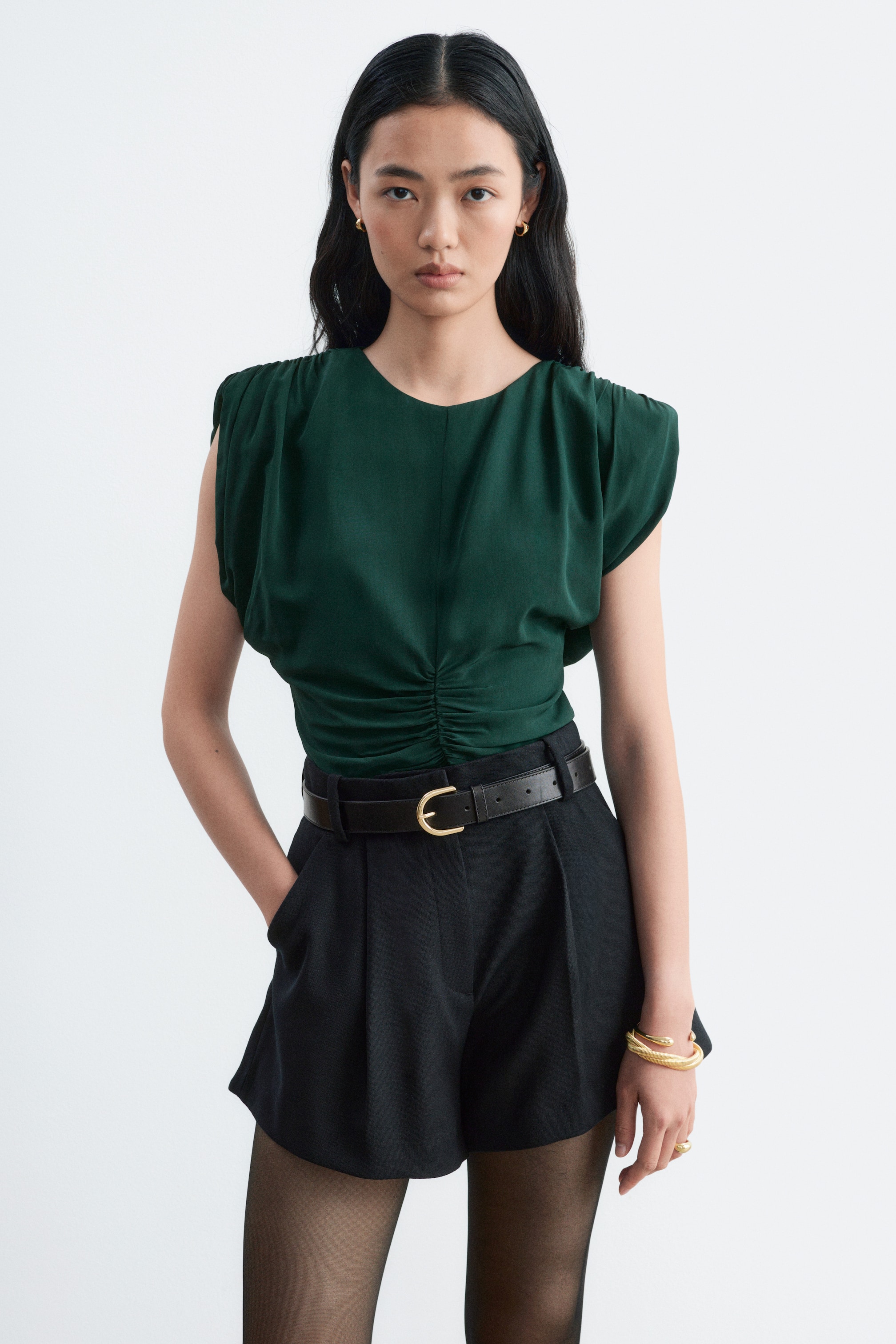Ver imagen más grande: La modelo lleva un top verde oscuro sin mangas, con fruncido en los hombros y en el centro delantero. El top está metido por dentro de unos pantalones cortos negros de cintura alta, combinados con un cinturón de cuero negro.