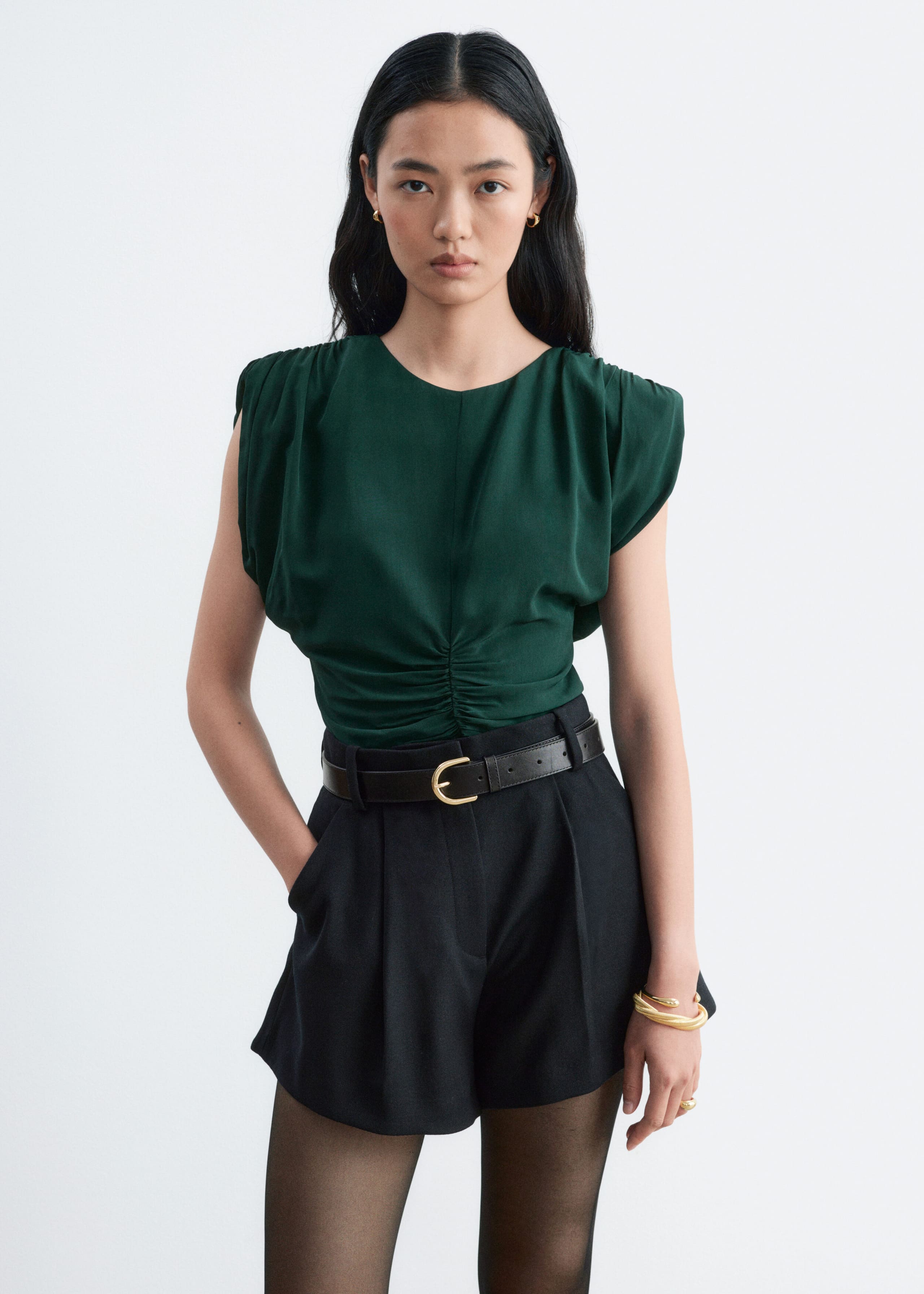 The model is wearing a dark green sleeveless top with ruching at the shoulders and center front. The top is tucked into high-waisted black shorts, styled with a black leather belt.