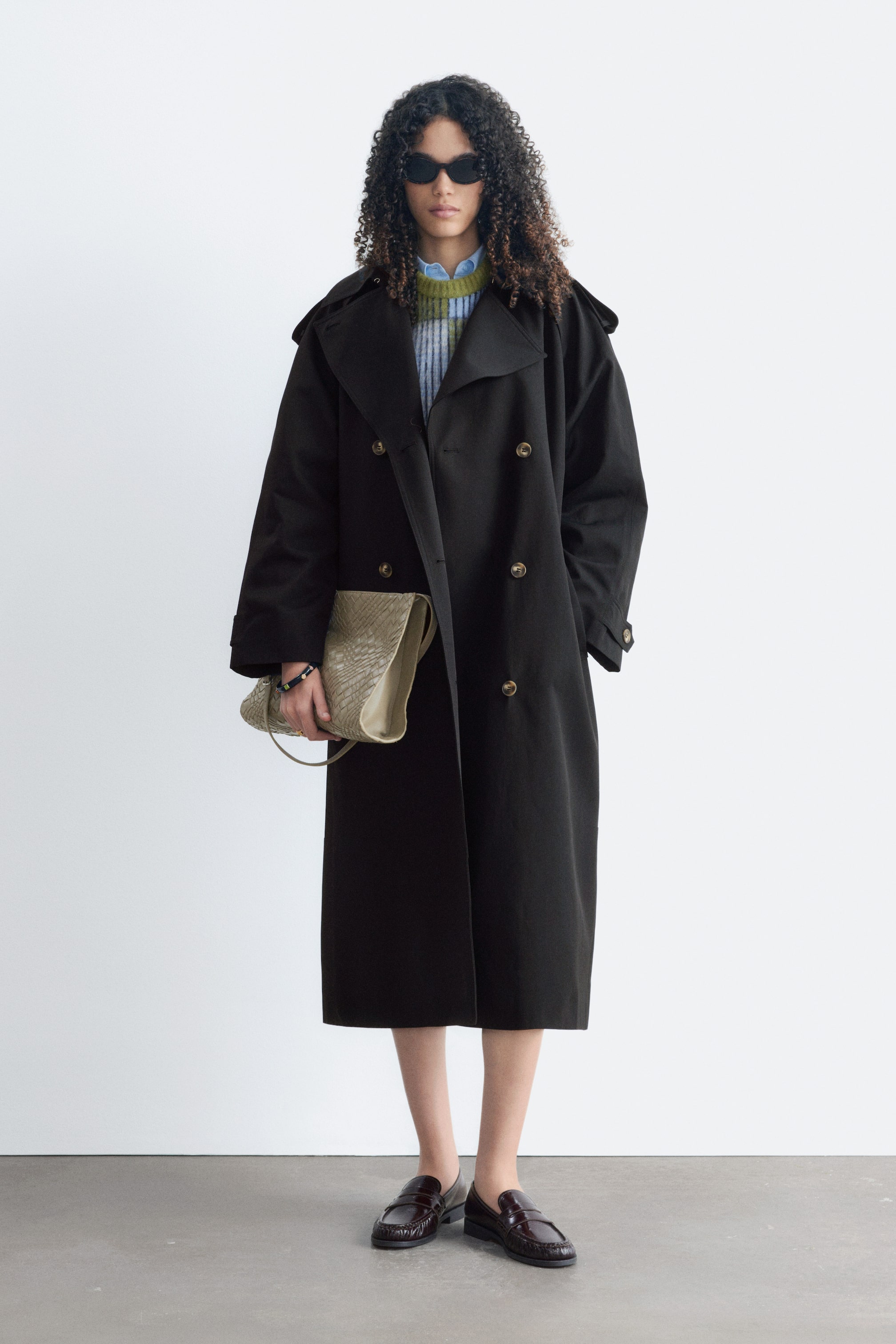 Größeres Bild ansehen: Ein schwarzer Trenchcoat in Relaxed Fit mit zweireihiger Front, Schulterklappen, Ärmellaschen und Schulterpasse wird vom Model offen getragen. Seine voluminöse Silhouette kommt deutlich zur Geltung, da er über einem gestreiften Pullover und einem Hemd getragen wird, während das Model eine geflochtene Tasche trägt.
