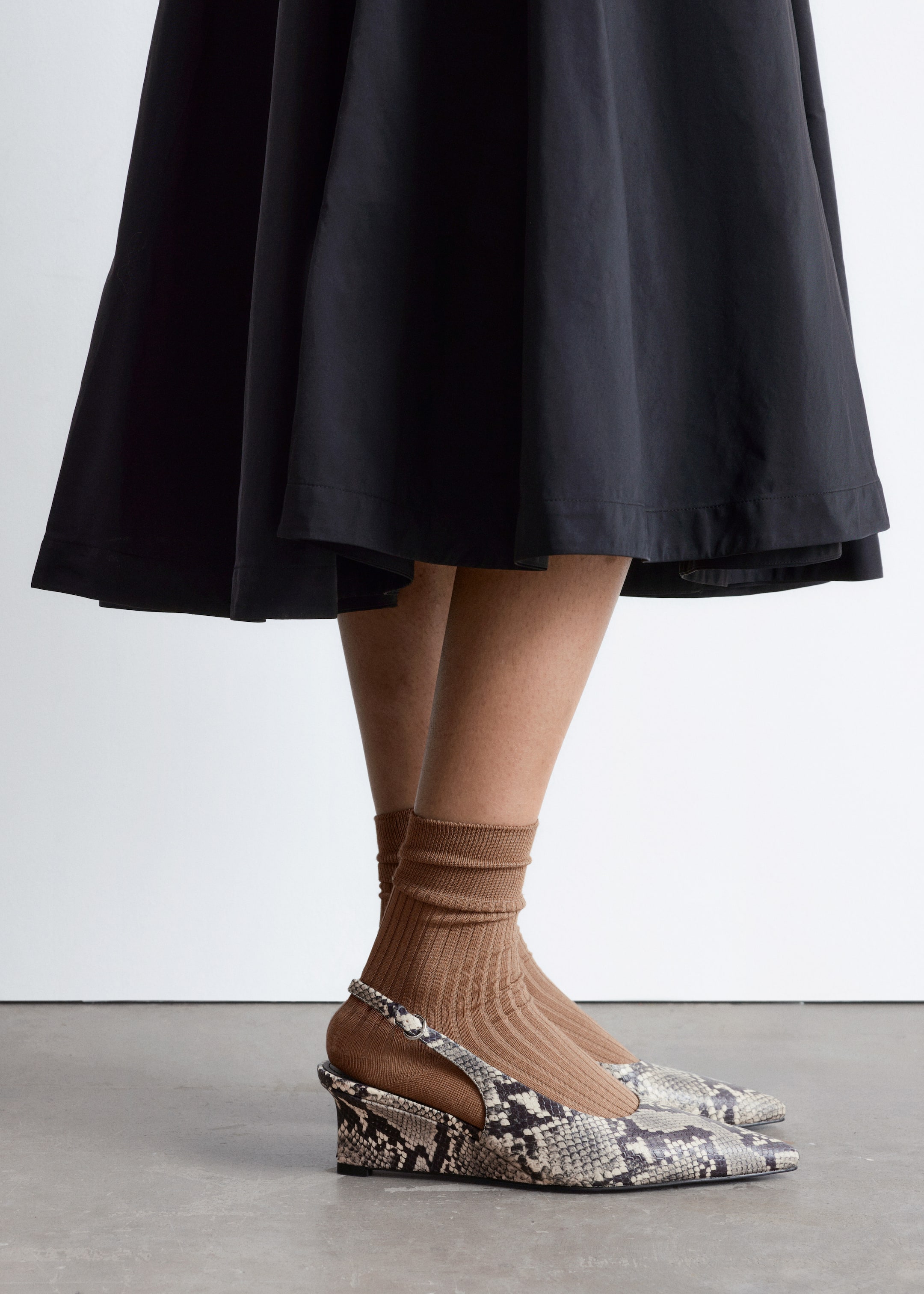 Größeres Bild ansehen: Ein schwarzer, weiter Rock fällt um die Beine. Braune, gerippte Socken sind an den Unterschenkeln sichtbar, kombiniert mit Keilschuhen im Schlangenhautmuster, die aus einem seitlich-vorderen Winkel zu sehen sind und eine spitze Zehenpartie sowie einen Riemen mit Schnalle aufweisen.