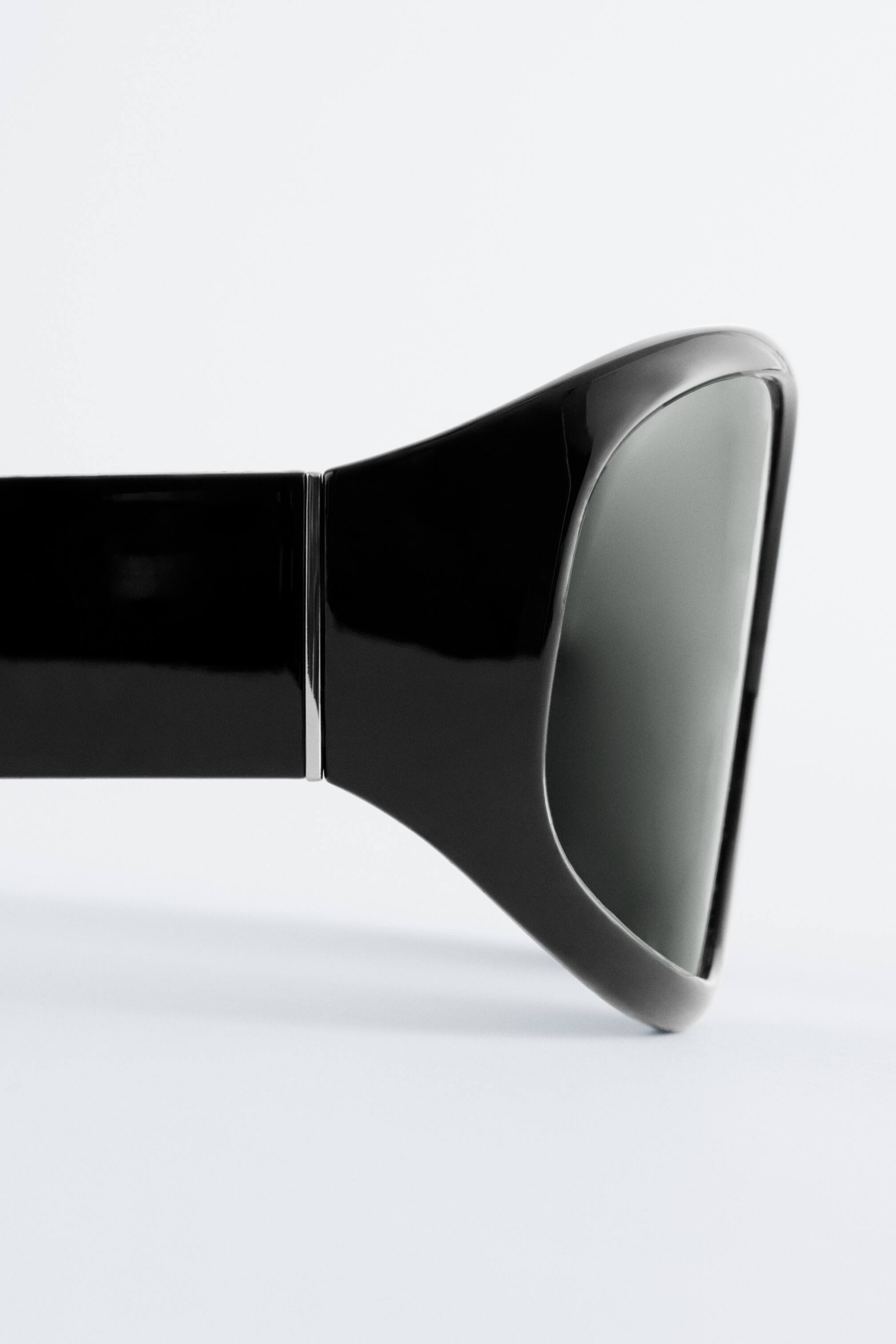 Zobrazit větší obrázek: Černé nadměrné čtvercové sluneční brýle jsou nakloněné doleva, odhalující lesklé, široké stranice, které se zužují směrem k kovovému detailu na pantu. Jemně zaoblený rámeček obklopuje tmavé, stínované čočky.