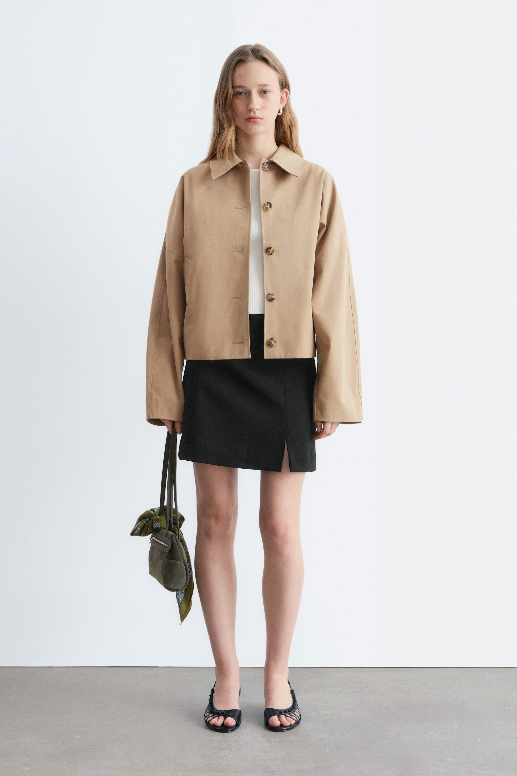 Agrandir l'image: Une veste courte beige présente un col à revers, six boutons marron sur le devant, et des manches longues amples, se portant de manière décontractée sur un haut blanc et se terminant au-dessus d'une jupe noire.