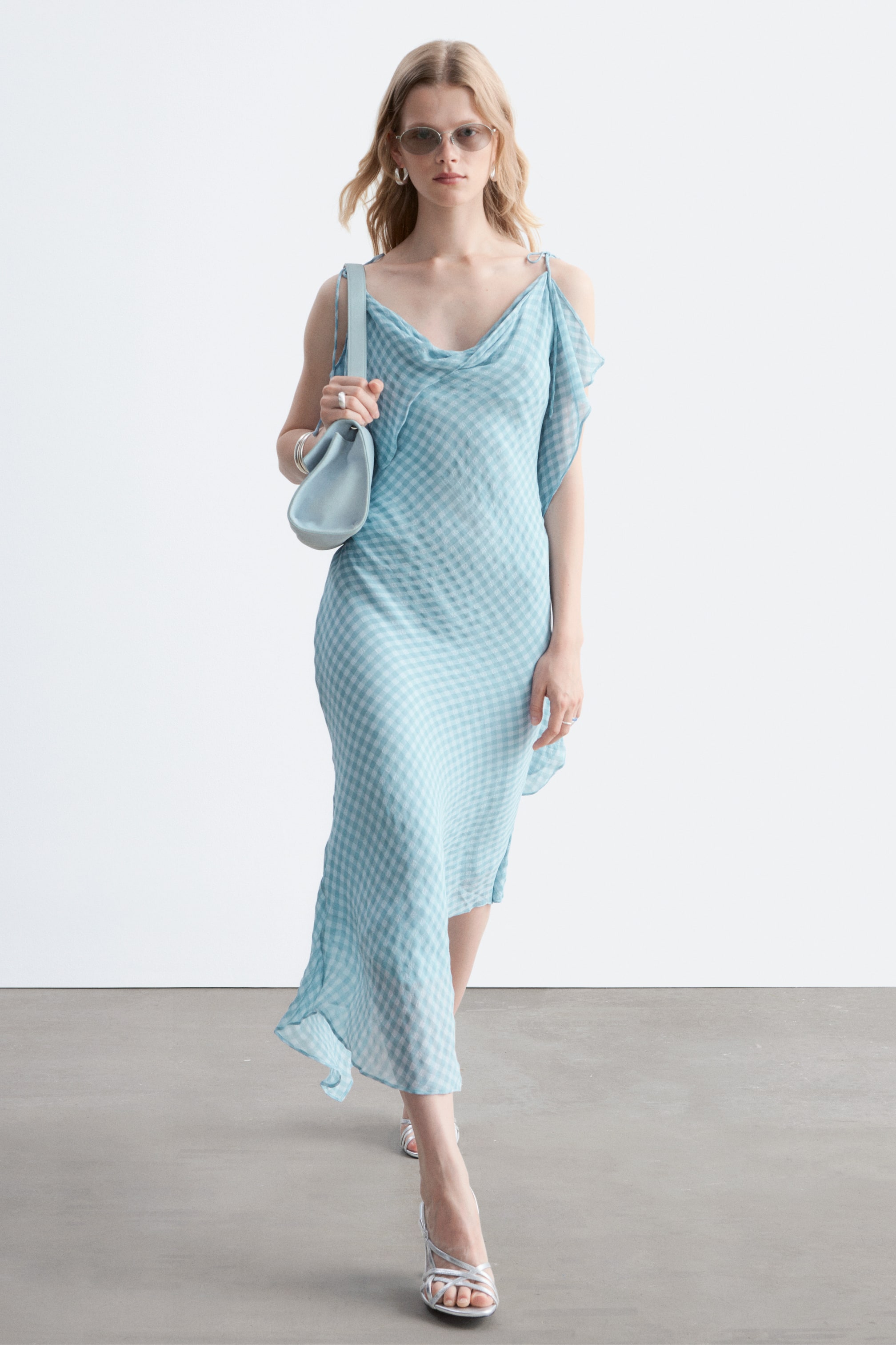 Ver imagen más grande: Un vestido midi de cuadros vichy en azul claro y blanco presenta un escote drapeado, finos tirantes y un detalle de hombro caído. Exhibe un dobladillo asimétrico y una silueta fluida, presentado desde un ángulo frontal.