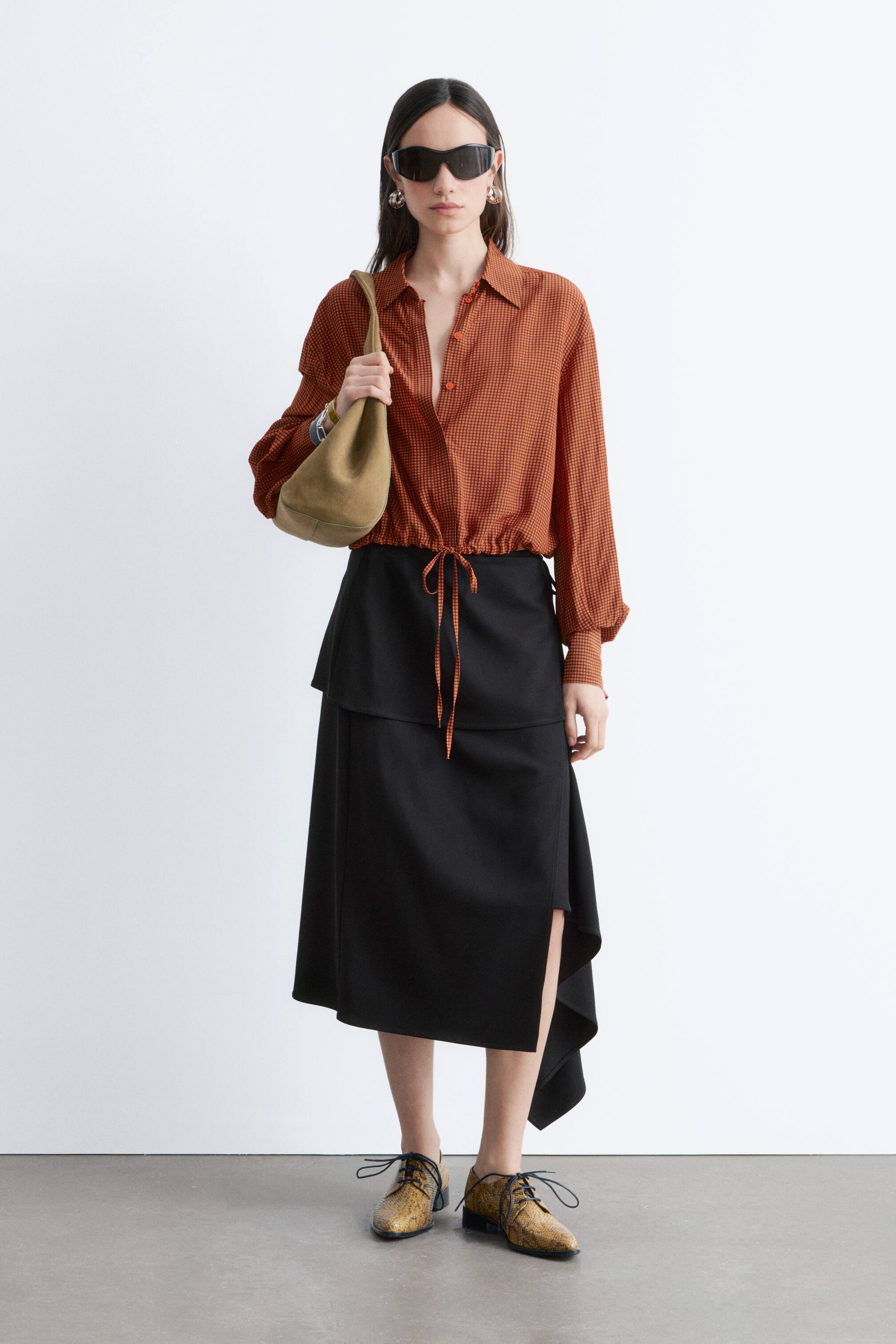 Visualizza immagine più grande: La modella indossa una camicia corta, caratterizzata da un motivo a micro quadretti arancioni e neri. La vita con coulisse è stretta e annodata, completata da un colletto classico, polsini con bottoni e un'abbottonatura nascosta.